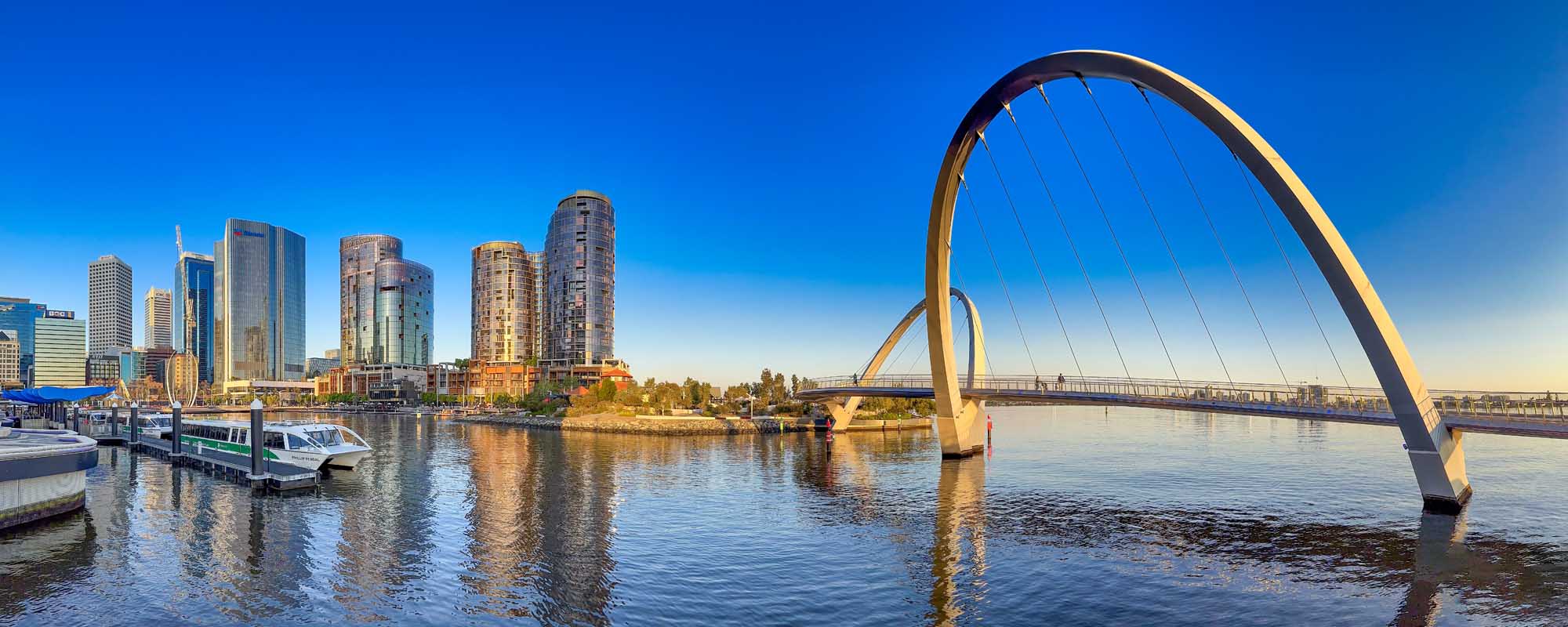 A panoramic waterfront view of Perth, Australia, featuring the iconic Elizabeth Quay Bridge with its distinctive curved arch design spanning the Swan River. The modern cityscape with high-rise buildings is reflected in the calm waters, with boats docked in the foreground and clear blue skies overhead.