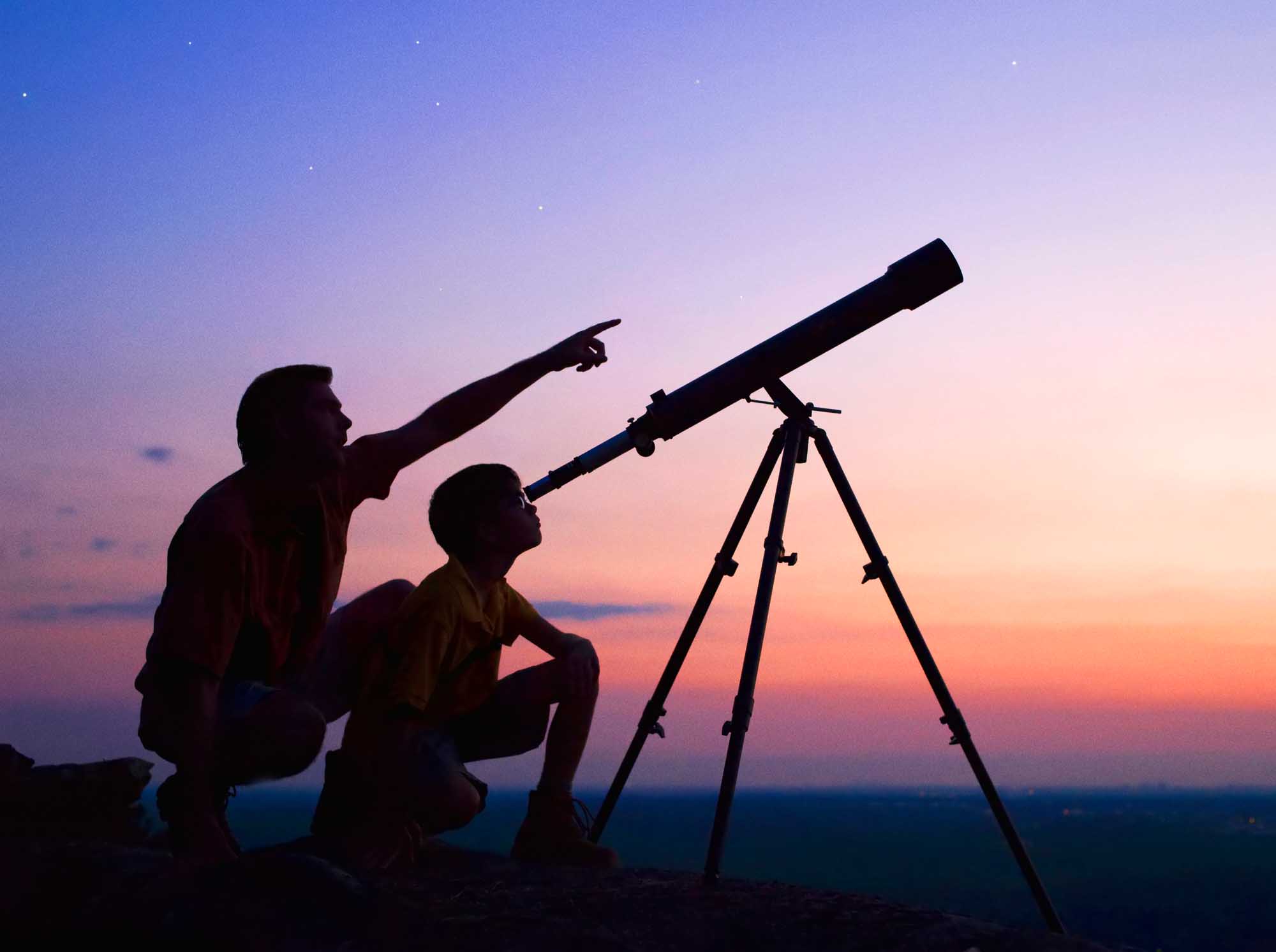 A silhouetted father and young son observe the night sky together through a telescope during a vibrant twilight hour. The scene captures a magical moment of discovery and shared wonder as stars begin to appear against a gradient sky transitioning from pink and orange hues to deep blue.