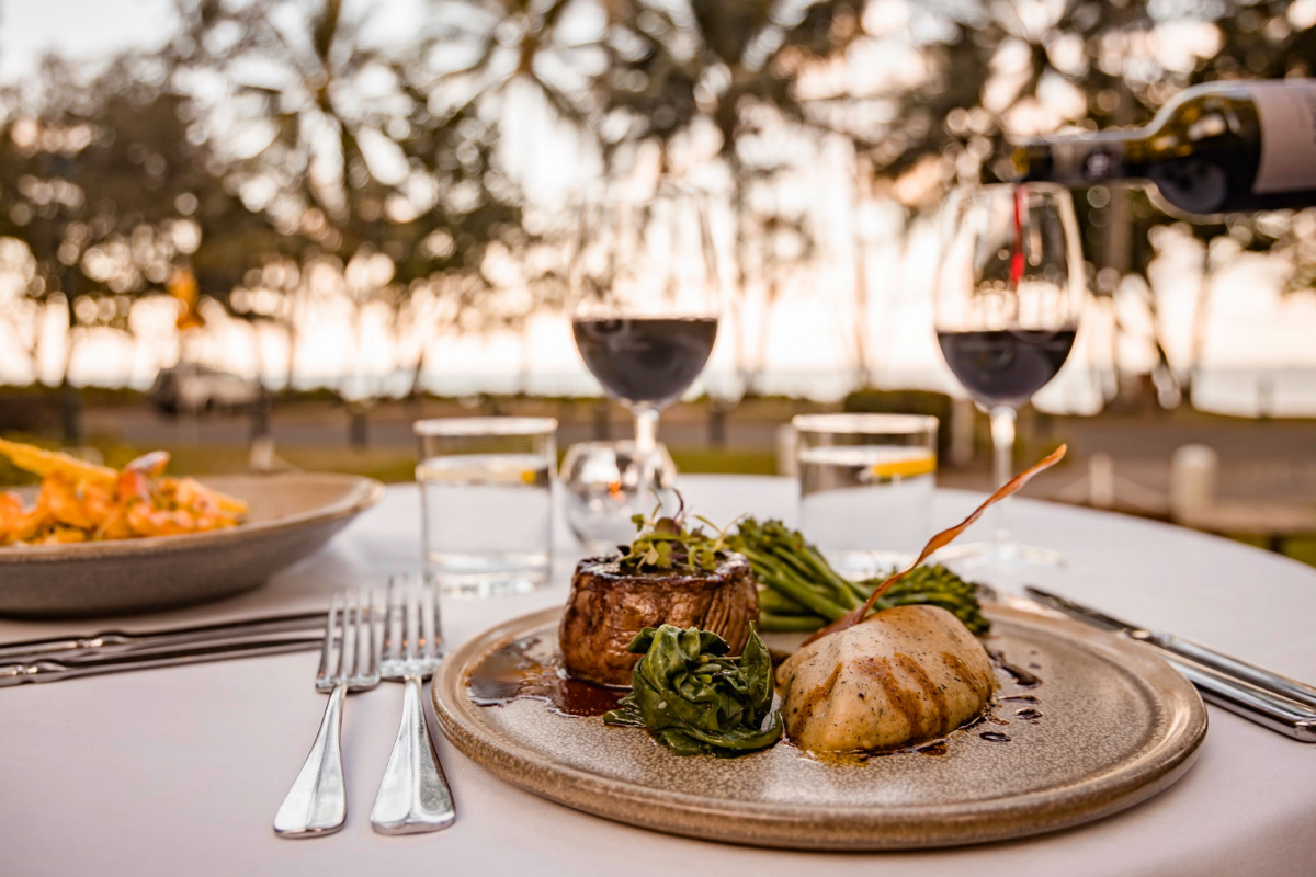 An elegant outdoor fine dining table setting at what appears to be an upscale resort or restaurant venue. The table features artfully plated food with fresh vegetables, seafood, and garnishes on a textured ceramic plate, paired with red wine in crystal glasses and formal place settings with white napkins and silverware. The background shows a blurred garden or vineyard setting with white umbrellas and additional diners, creating an atmosphere of refined hospitality and culinary excellence.