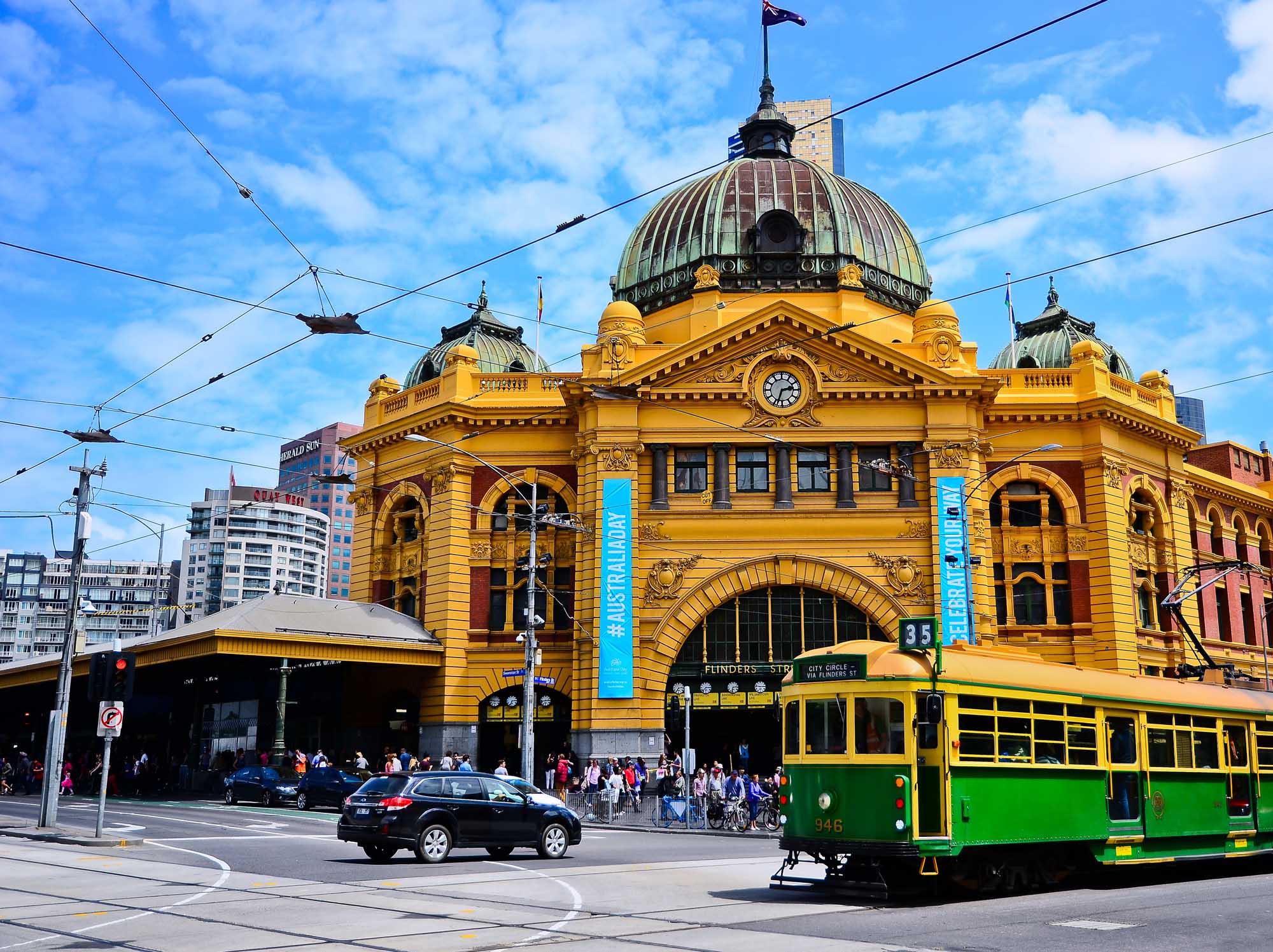A vibrant daytime view of Melbourne's iconic Flinders Street Station, an ornate yellow Victorian-era building with a distinctive dome and clock tower, featuring a vintage green and yellow tram (number 35) in the foreground. The architectural landmark is a prominent transport hub in the city centre, with overhead tram wires and modern urban buildings visible in the background under blue skies.