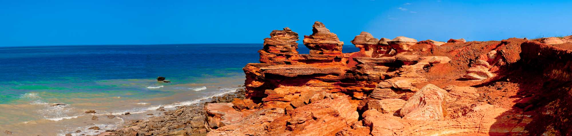 Dramatic red sandstone cliffs tower above the pristine waters at Gantheaume Point, located at the southern end of Cable Beach in Broome, Western Australia. The vibrant rust-colored rock formations meet turquoise ocean waters under a brilliant blue sky, creating a striking natural landscape.