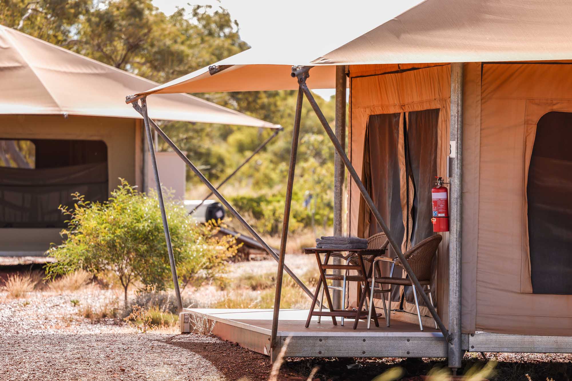 A luxurious glamping tent setup featuring an orange canvas structure with an extended awning, wooden deck platform, and comfortable seating area. The scene showcases a fire extinguisher mounted on the tent and demonstrates upscale outdoor accommodation amenities in a natural, arid landscape with scattered vegetation and trees.