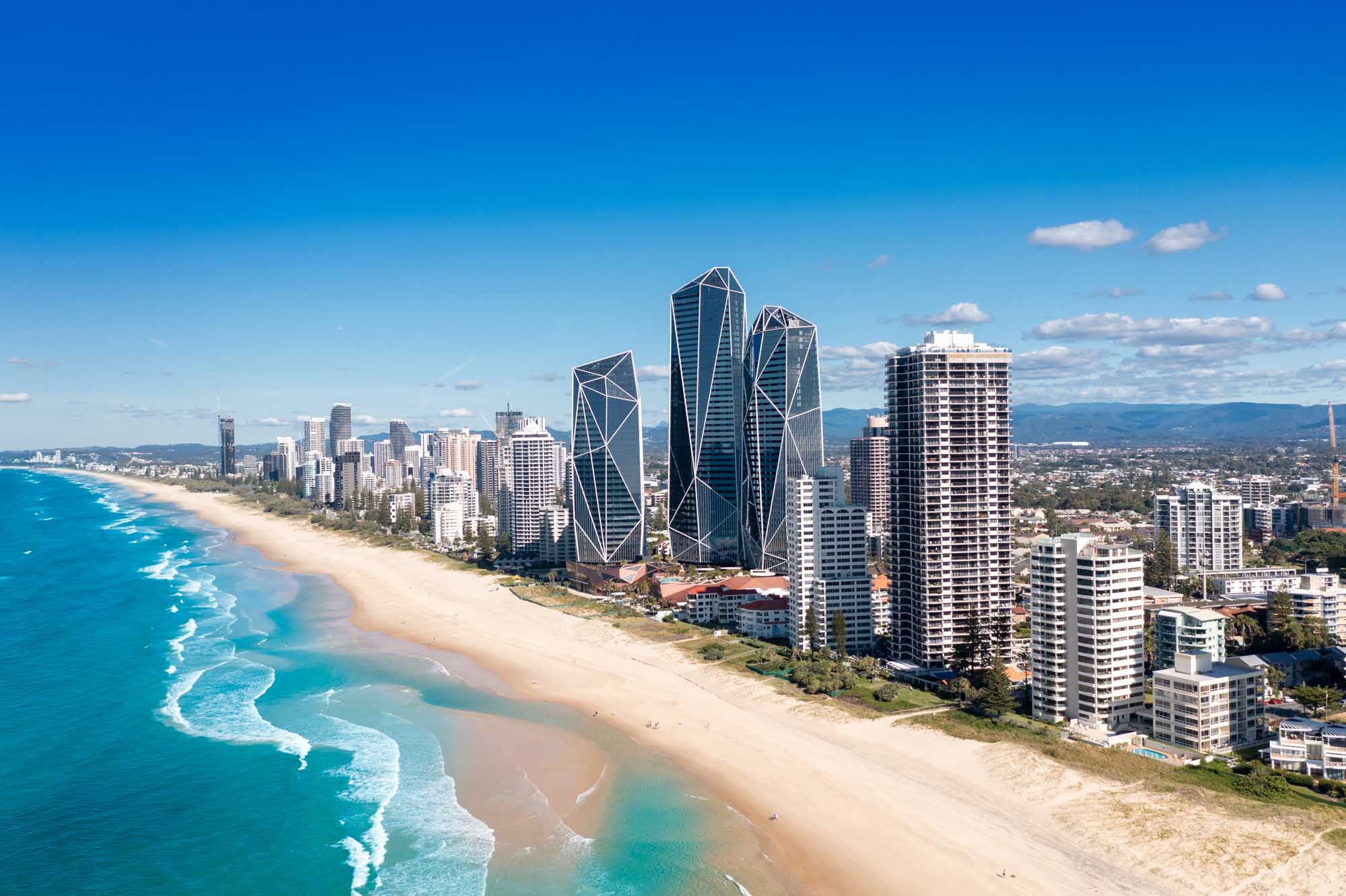 An aerial photograph of Australia's Gold Coast skyline featuring distinctive modern high-rise towers, pristine sandy beaches, and turquoise ocean waters. The image captures the coastal cityscape with mountains visible in the distance under clear blue skies.