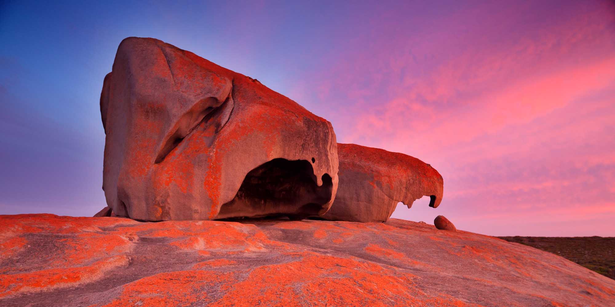 Massive granite rock formations illuminate in vivid pink and orange hues during sunset on Kangaroo Island, South Australia. The distinctive boulder formations, known as the Granite Guardians, create dramatic silhouettes against a colorful twilight sky with purple and pink gradients.
