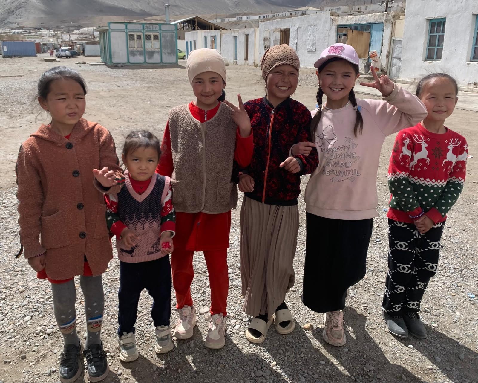 A cheerful group of six children standing together in an arid, mountainous settlement with simple