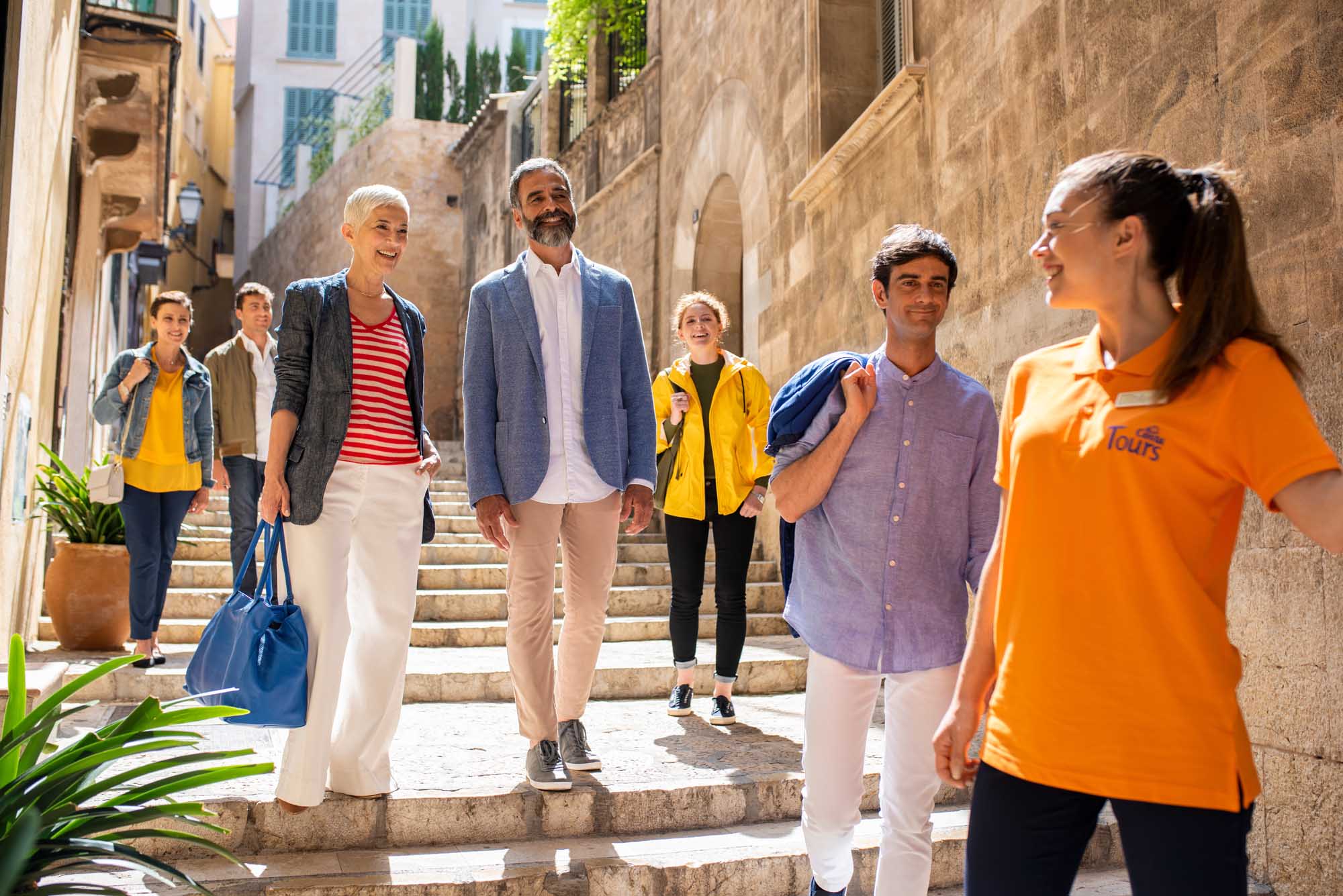 A diverse group of tourists walks through a charming narrow stone alleyway in a historic Mediterranean town, led by a tour guide in an orange branded polo shirt. The scene captures the essence of cultural tourism, with colorful casual clothing and a mix of ages enjoying a guided walking tour through traditional architecture.