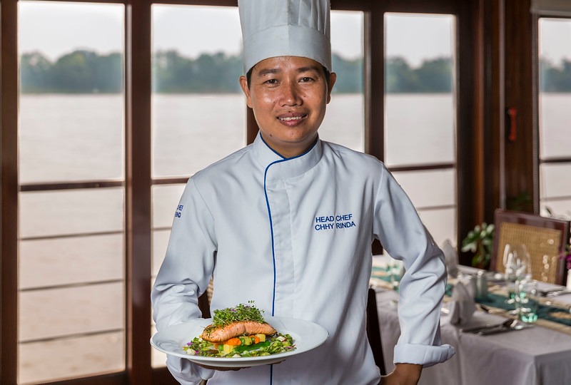 A professional head chef in white chef's coat and tall hat stands smiling while holding a beautifully plated dish with salmon, greens, and garnish. The setting is an elegant dining room aboard a river cruise vessel, with large windows overlooking a calm river and distant landscape, suggesting a Southeast Asian river cruise destination.