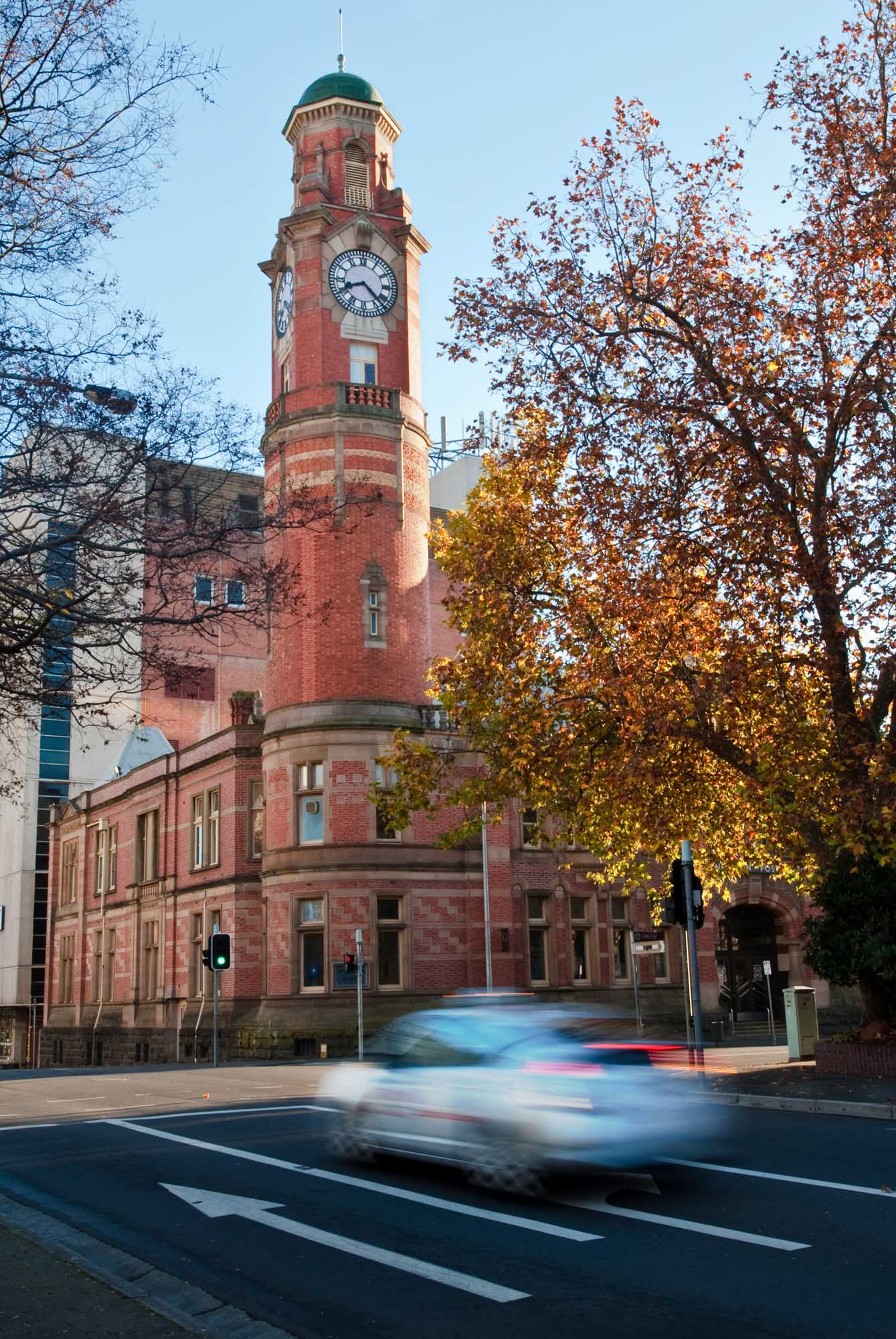 A striking red brick Victorian-era clock tower with a green domed roof stands prominently in an urban square, framed by autumn foliage. A blurred taxi passes through the foreground at traffic lights, capturing the dynamic energy of this historic Australian landmark during an early morning autumn scene.