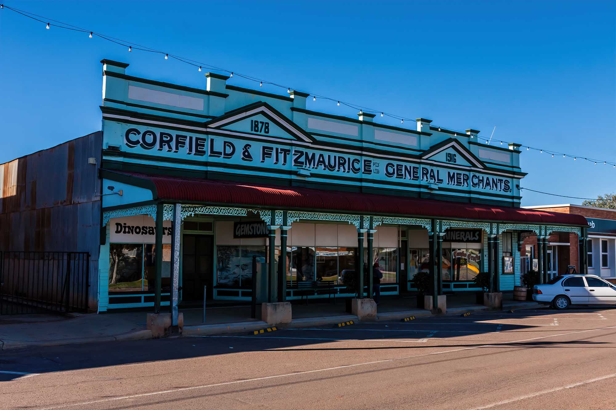 A beautifully preserved heritage storefront in Winton, Australia, featuring the distinctive turquoise and black façade of the Corfield & Fitzmaurice General Merchants building dated 1878-1916. The building showcases classic Australian colonial architecture with decorative ironwork, large display windows, and period signage.