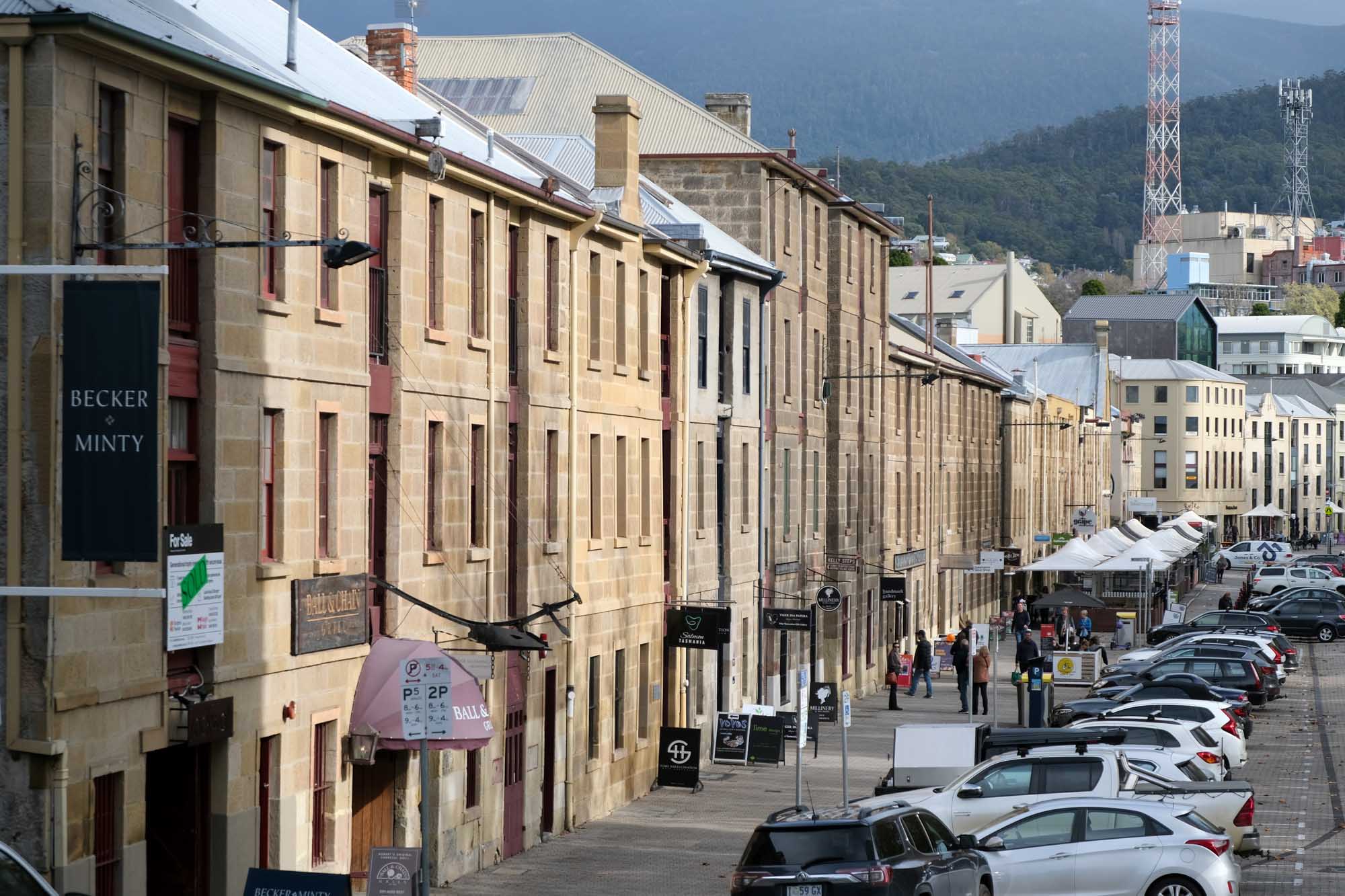 A picturesque row of heritage sandstone buildings lines a pedestrian plaza at Salamanca Place in Hobart, Tasmania. The formerly utilitarian port warehouses now house restaurants, galleries, craft shops, and offices, with visitors strolling the cobblestone street beneath mountains in the background.