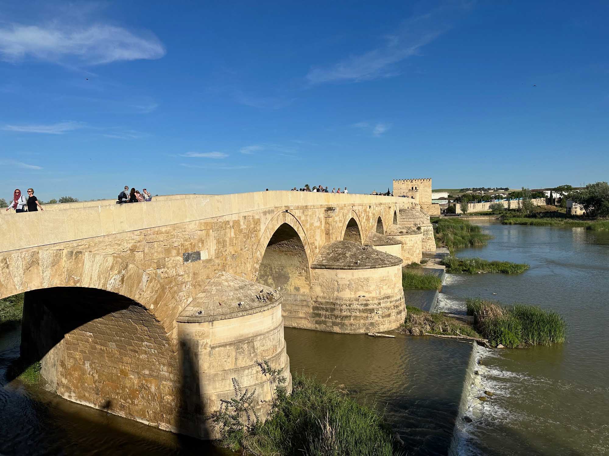 A beautifully preserved medieval stone bridge with multiple arches spans across a calm river, featuring characteristic Romanesque architecture with cylindrical defensive towers. The bridge is lined with visitors enjoying the scenic views, with a fortified tower visible on the right bank and lush green vegetation framing the waterway under a clear blue sky.