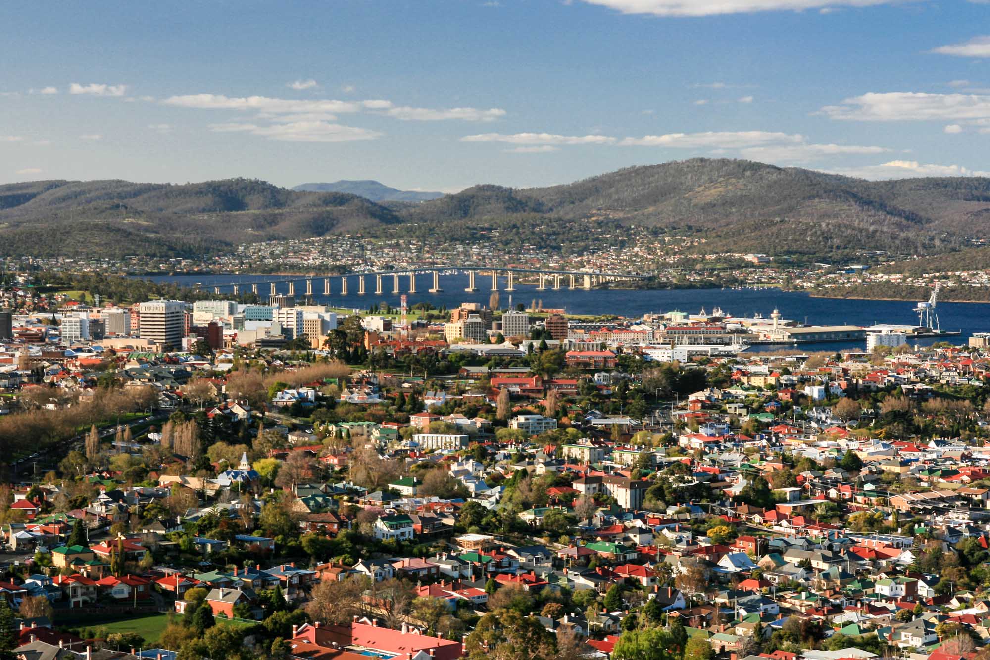 A panoramic aerial view of Hobart, Tasmania, showing the downtown skyline, residential neighborhoods, and the iconic Tasman Bridge spanning the blue Derwent River. Forested hills and mountains surround the city under a clear sky with scattered clouds.