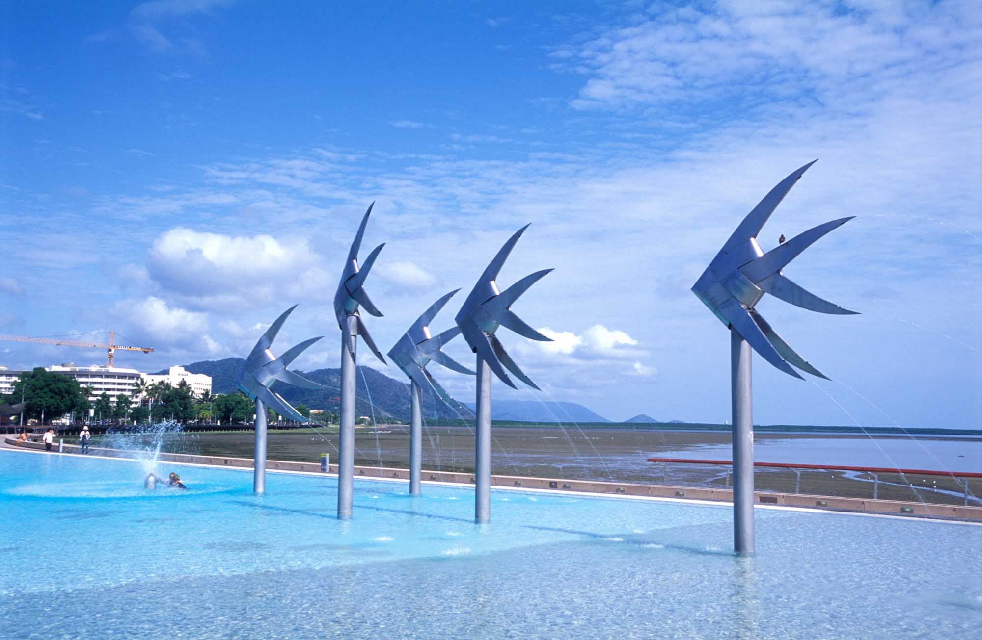 A striking public swimming pool in Cairns, Australia, featuring distinctive blue pinwheel wind sculptures rising from the water. The pool overlooks the tropical coastline with mountains visible in the distance, set against a bright blue sky with scattered clouds.