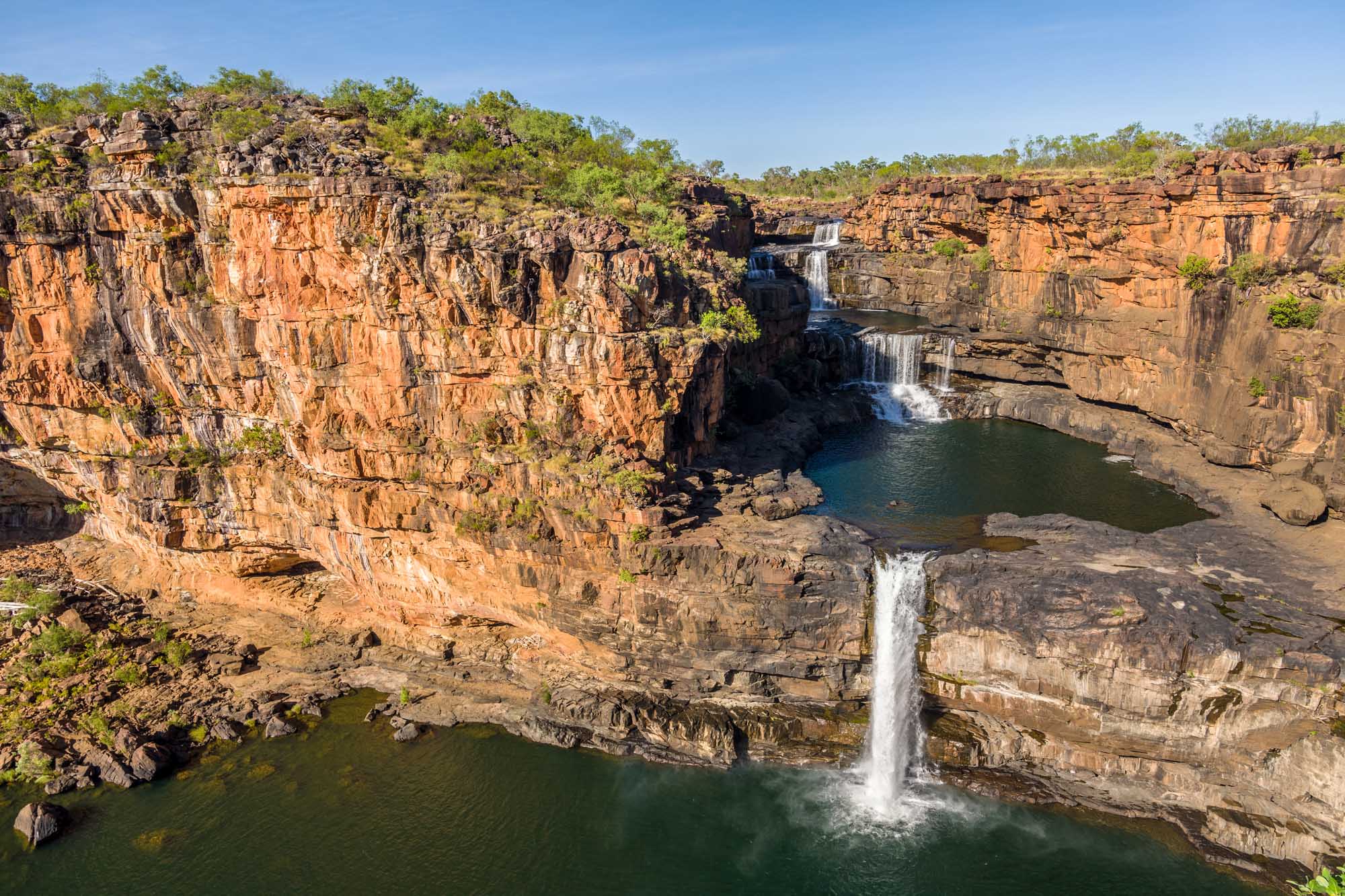 A dramatic waterfall cascades into a pristine turquoise pool surrounded by towering red sandstone cliffs in Kakadu National Park, Western Australia. The rugged landscape features layered rock formations and sparse vegetation characteristic of Australia's Top End.