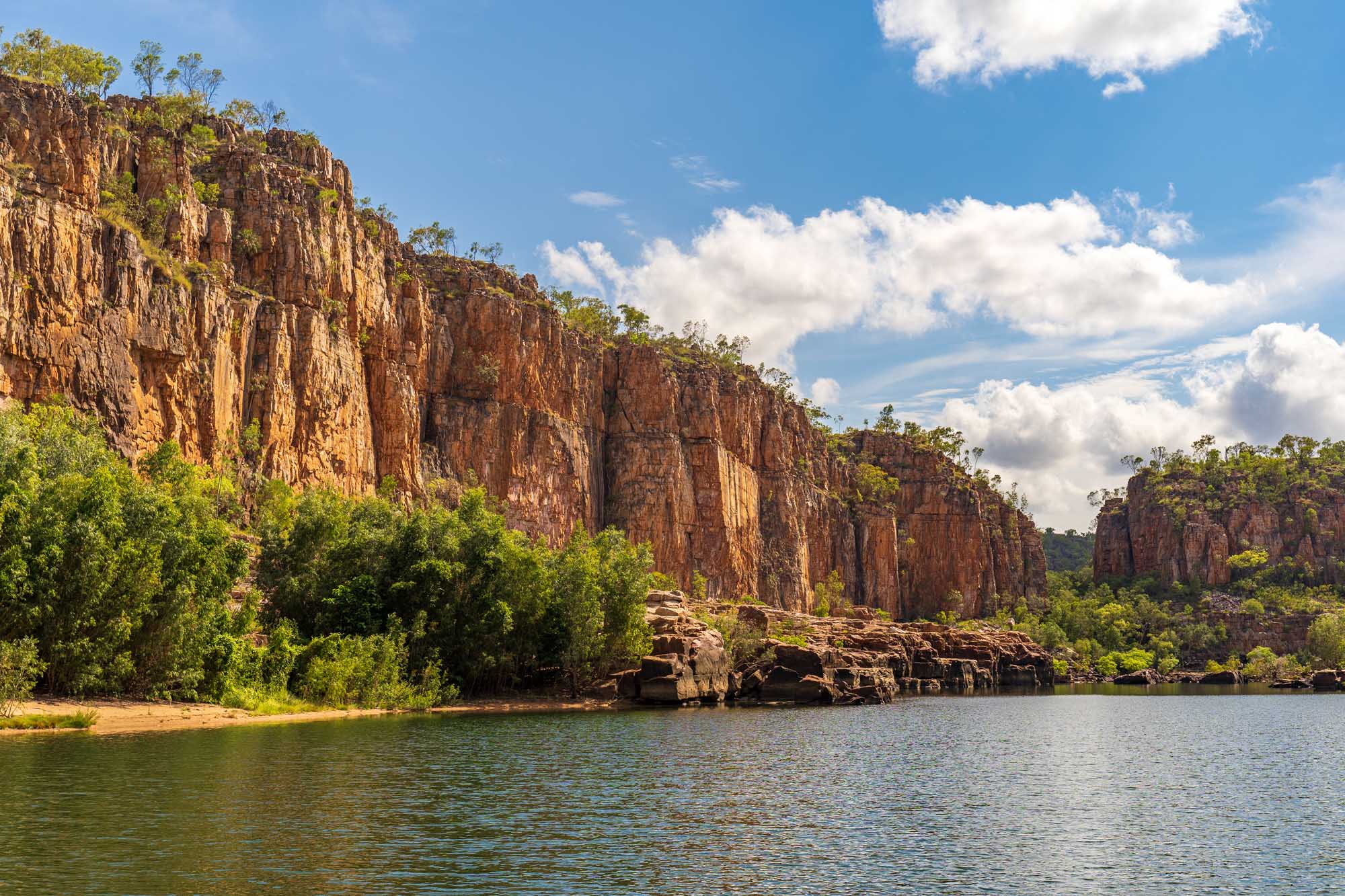 Dramatic red sandstone cliffs rise majestically from the tranquil waters of Katherine Gorge in Nitmiluk National Park, Northern Territory, Australia. Lush green vegetation clings to the cliff faces under a brilliant blue sky with white clouds, creating a stunning natural landscape ideal for river cruises and adventure tourism.