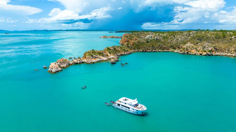 A small expedition cruise vessel anchors in brilliant turquoise waters near rocky, vegetation-covered coastline, characteristic of Australia's remote Kimberley region. The pristine tropical seascape features dramatic limestone formations and lush greenery under a partly cloudy sky.