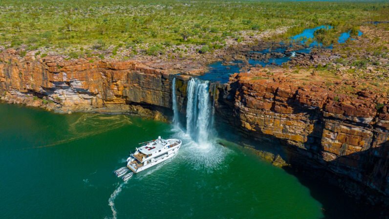 An expedition cruise vessel navigates beneath a dramatic waterfall cascading from ancient red rock cliffs into a pristine emerald pool, surrounded by rugged tropical landscape in Australia's remote Kimberley region.