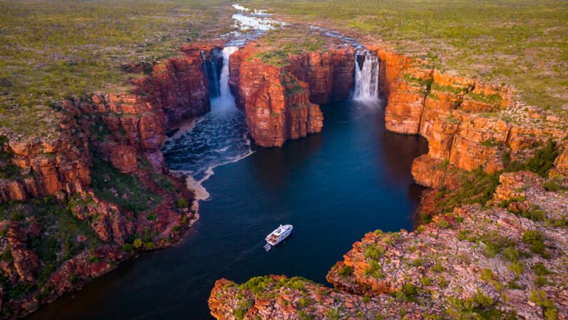 An aerial view of the spectacular King George Falls in Western Australia's remote Kimberley region, with the Kimberley Quest expedition vessel anchored in the deep pool below the twin cascading waterfalls. The dramatic rust-red cliff faces frame the pristine blue waters, surrounded by sparse vegetation typical of the Australian outback.