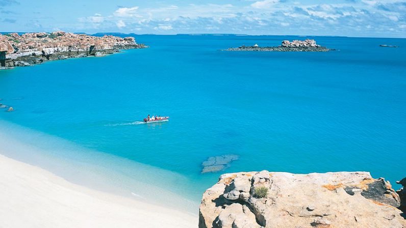 A small expedition vessel navigates the crystalline turquoise waters of Vansittart Bay, with dramatic rocky outcrops and pristine white sand beaches visible in the foreground and surrounding coastline dotted with weathered sandstone formations.