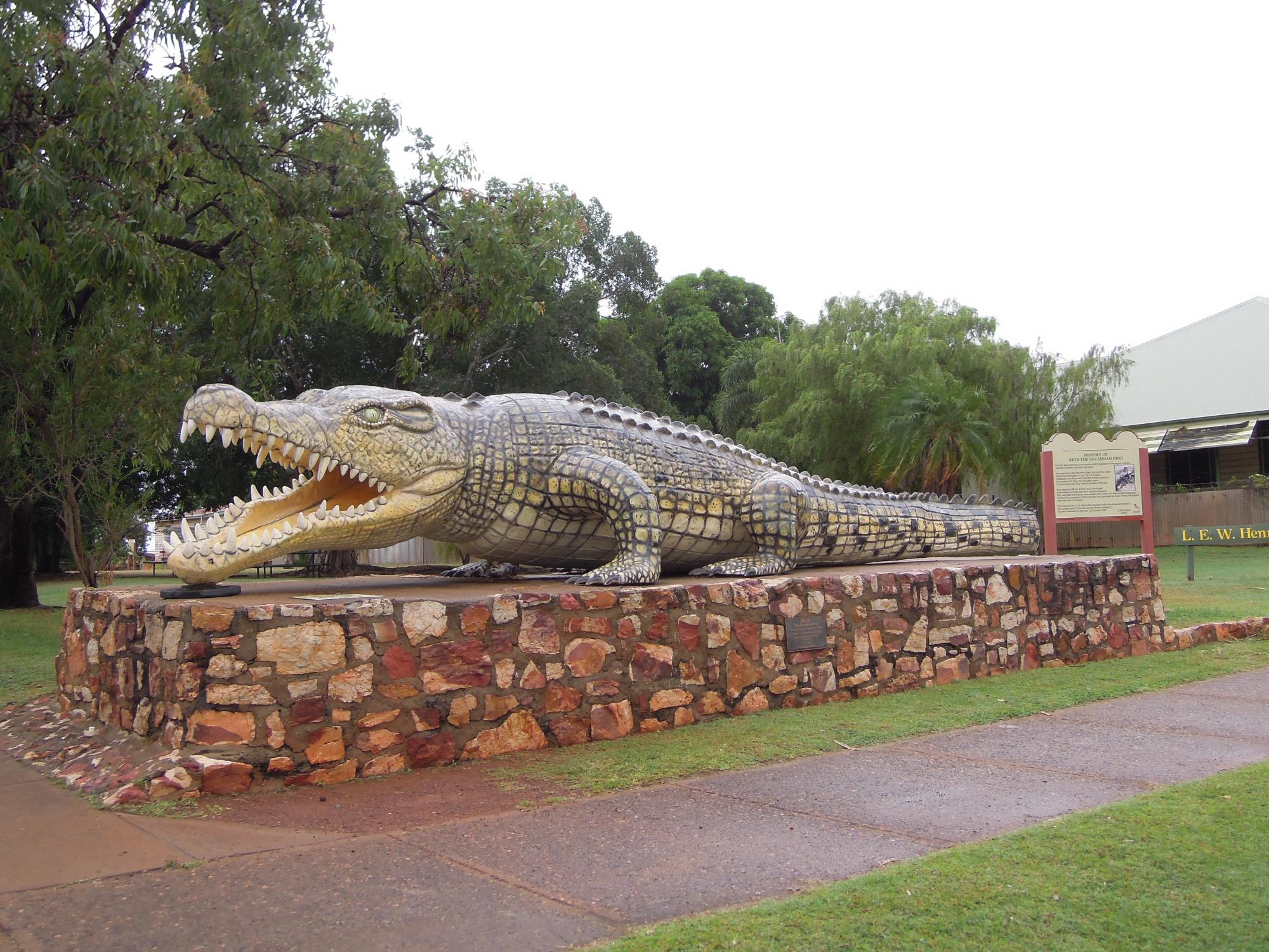 A large concrete crocodile sculpture displayed on a stone base in Normanton, featuring detailed carved teeth and scales. The statue is positioned as a roadside attraction with informational signage and is surrounded by native Australian trees and landscaping.