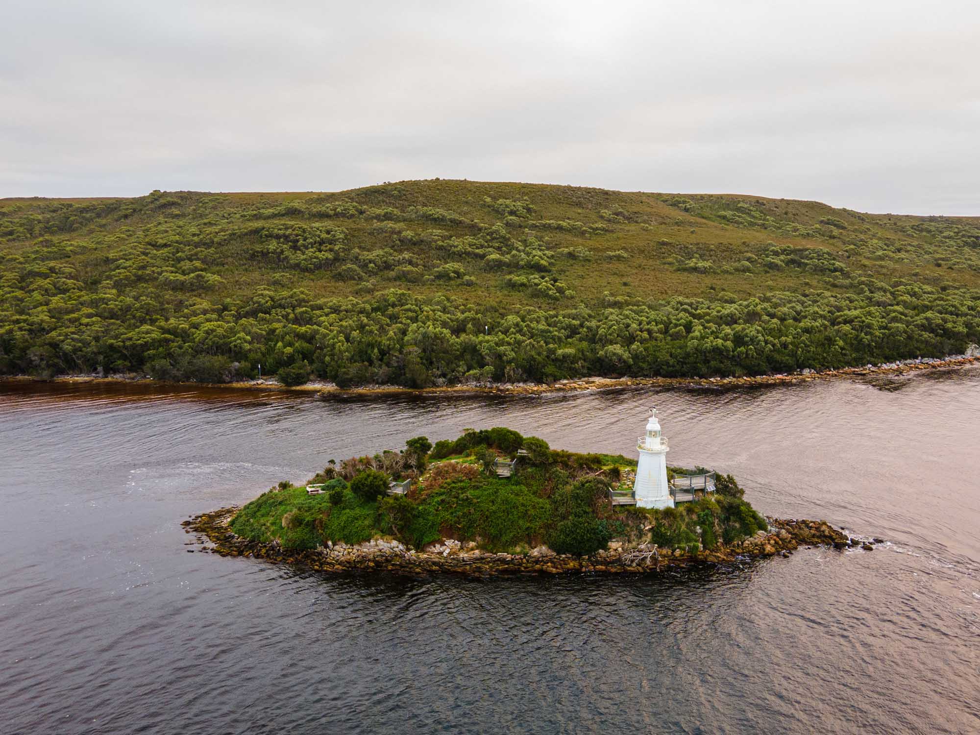 A white lighthouse stands on a small, densely vegetated island in Macquarie Harbour, Tasmania, surrounded by calm waters with forested hillsides visible in the background. This is a scenic view of Hell's Gates, a notable maritime landmark in Tasmania's southwest.