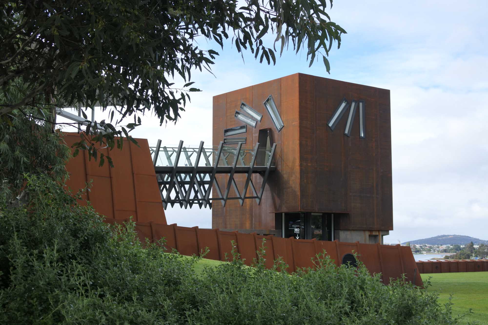 The striking rust-colored exterior of MONA (Museum of Old and New Art) in Hobart, Tasmania, featuring distinctive architectural elements including protruding metal tubes and angular geometric forms. The avant-garde museum building is set against a backdrop of water and distant hills, with native vegetation in the foreground.