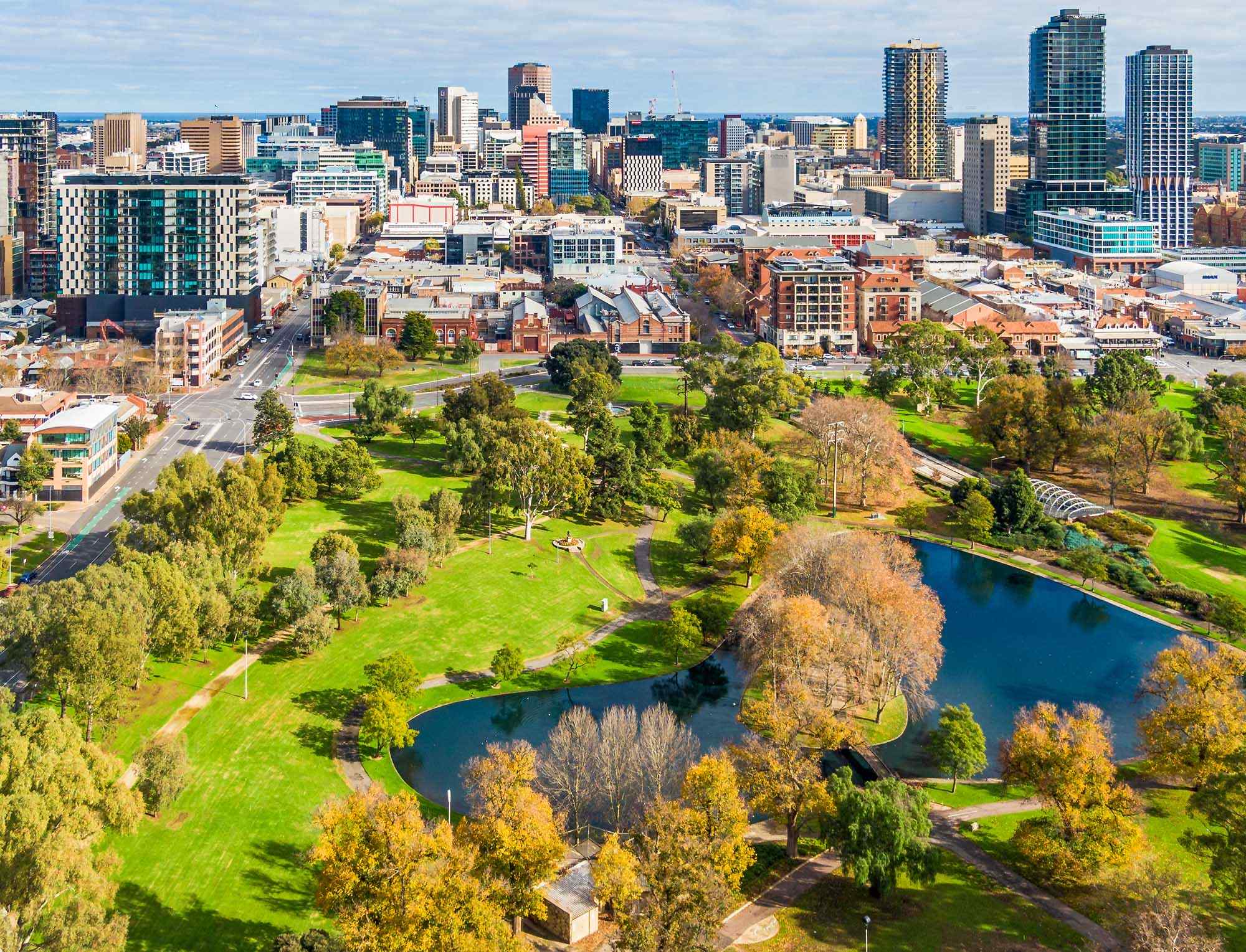 An aerial view of Melbourne's central business district featuring modern skyscrapers and historic architecture, with a lush green park and meandering Yarra River in the foreground. The image captures the contrast between urban development and natural green spaces during autumn, with clear blue skies overhead.
