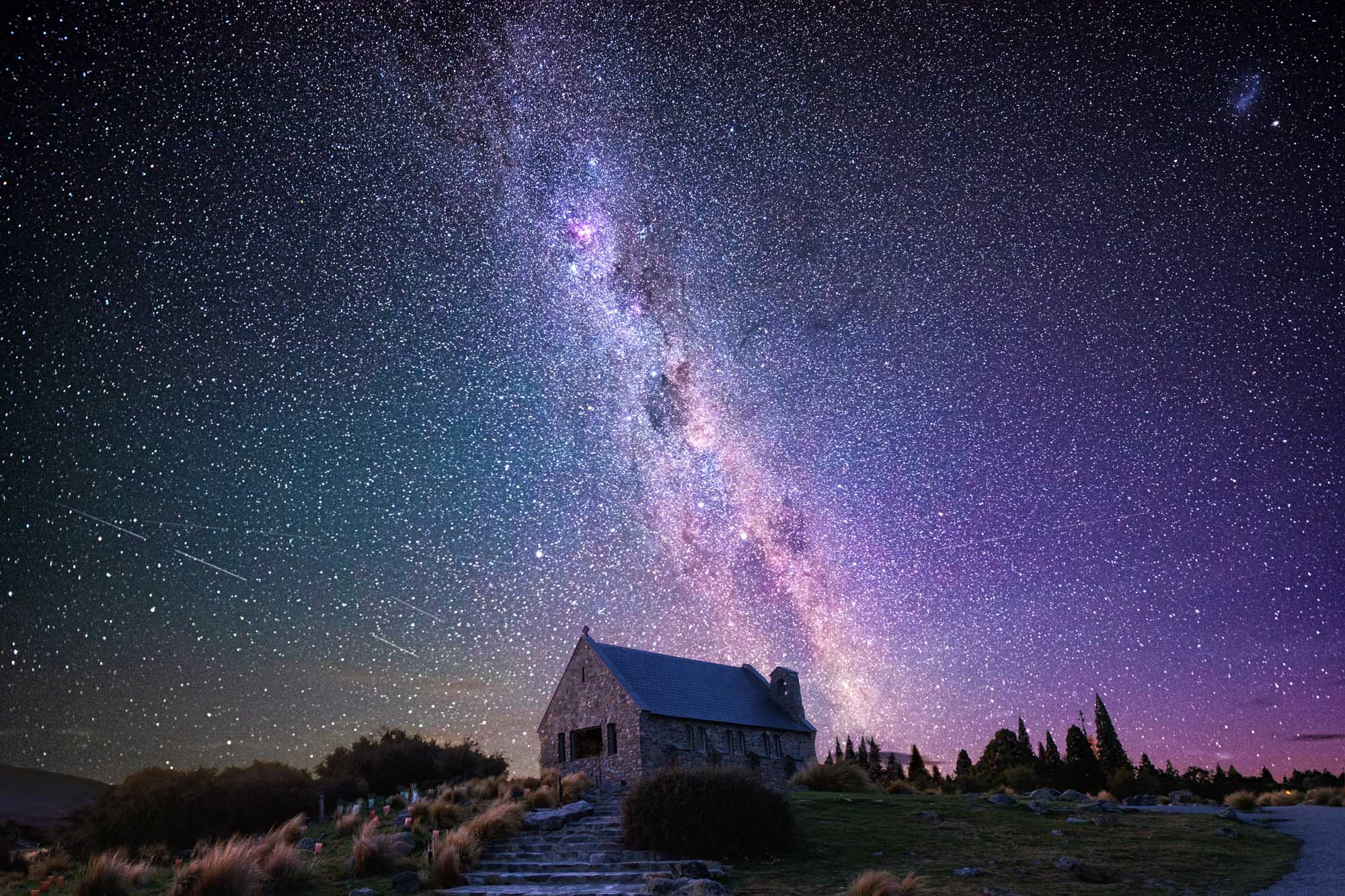 A breathtaking nighttime photograph of the iconic Good Shepherd's Church silhouetted against a stunning display of the Milky Way galaxy spanning across the sky in vibrant purples, blues, and pinks. The stone chapel sits on a hillside surrounded by native vegetation, with a meteor streak visible on the left side of the frame, showcasing New Zealand's exceptional stargazing conditions.
