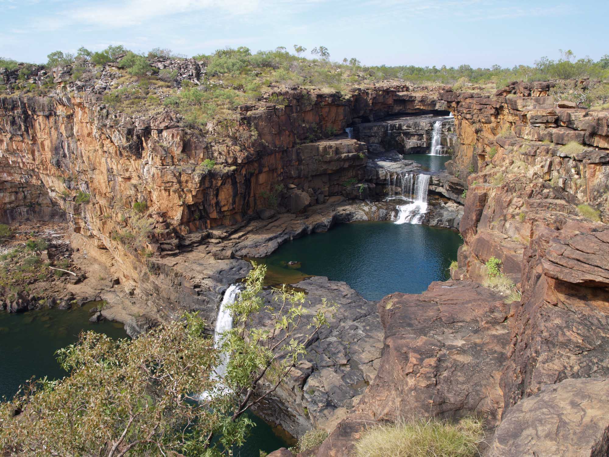 A stunning natural waterfall landscape in Western Australia's Kimberley region, featuring multi-tiered cascades flowing into pristine emerald pools surrounded by dramatic red sandstone cliffs and sparse spinifex vegetation characteristic of the remote Australian outback.