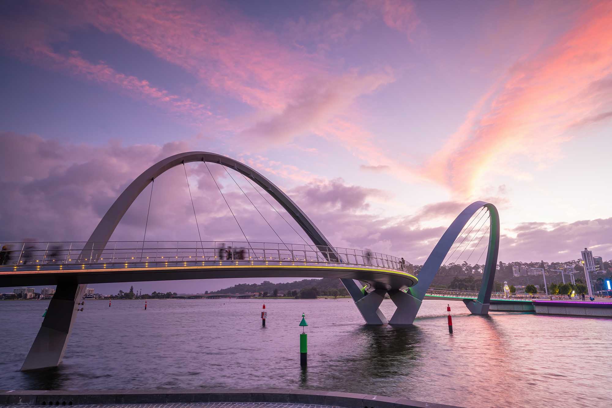 A striking modern arch bridge spans across a river at golden hour, with its distinctive curved design illuminated against a vibrant purple and pink sunset sky. The architectural landmark features elegant cable stays and is reflected in the calm water below, with the Perth city skyline visible in the background.