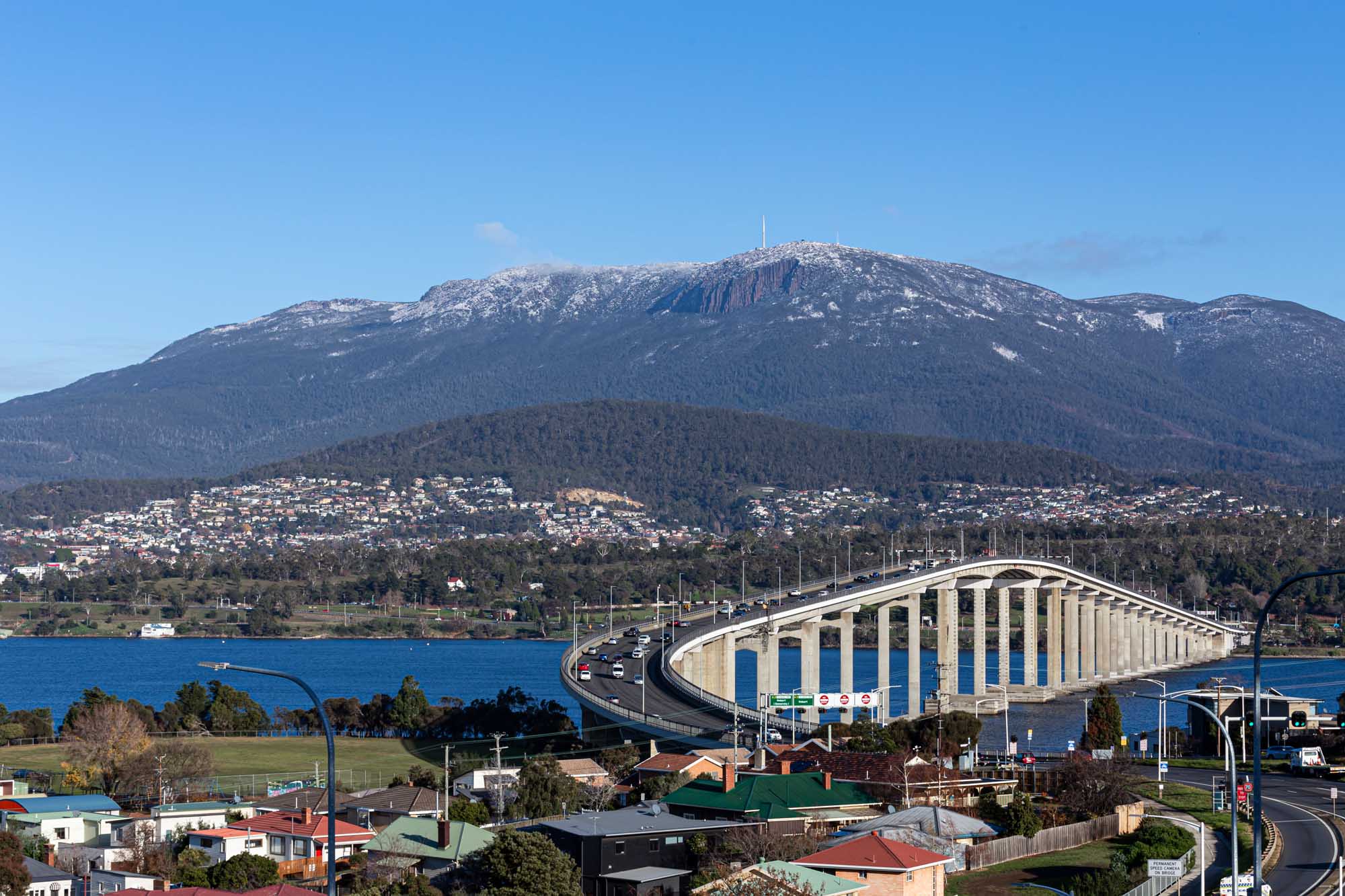 A scenic view of Hobart, Tasmania, featuring the iconic Tasman Bridge spanning the Derwent River with Mount Wellington snow-capped in the background. Residential neighborhoods and forested slopes line the shores beneath clear blue skies.