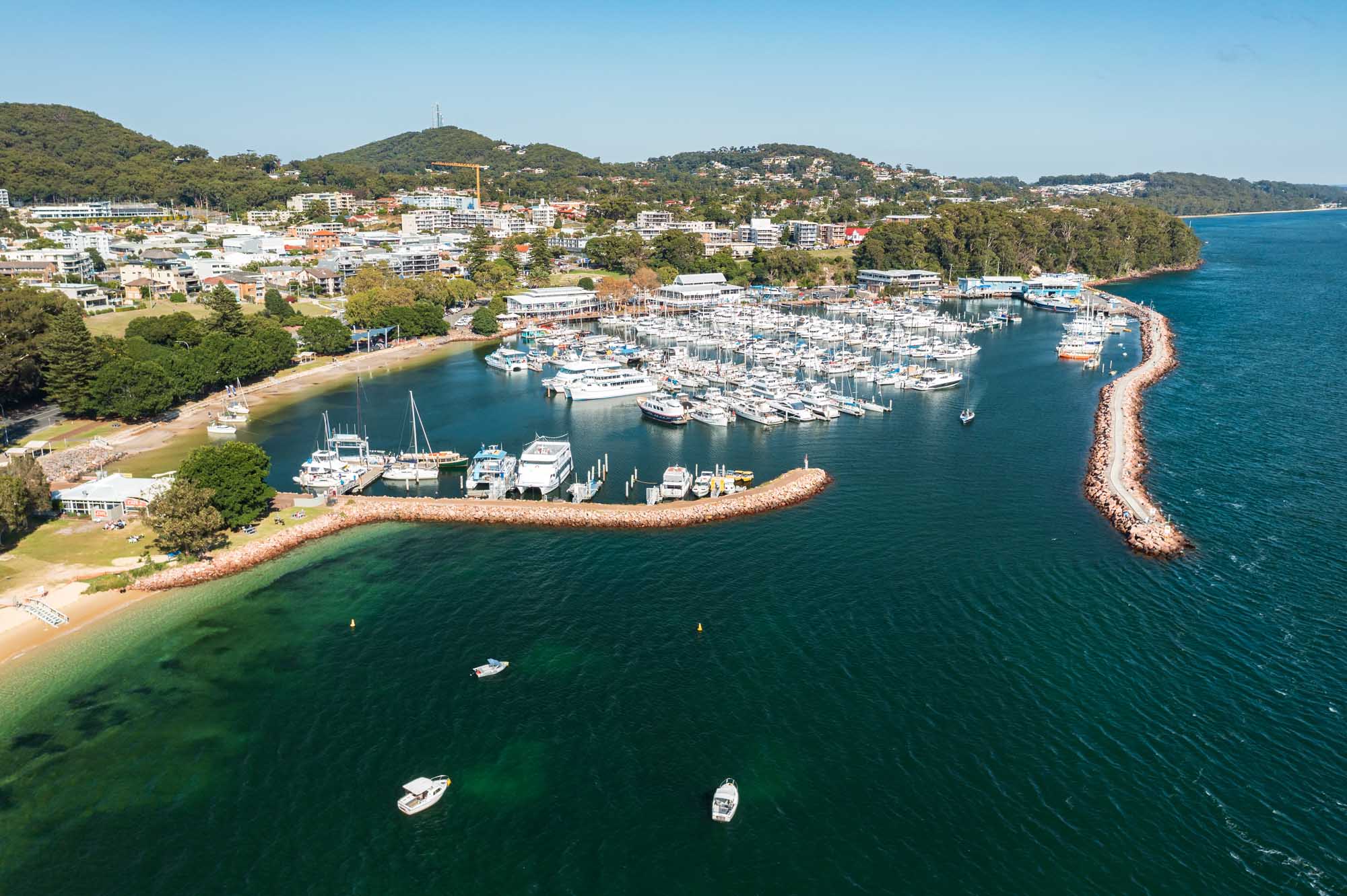 An aerial photograph of Nelson Bay marina in Port Stephens, NSW, Australia, showcasing a densely packed marina filled with white boats, distinctive red rock breakwaters, sandy beaches, and the surrounding coastal town nestled against forested hills with clear turquoise waters.