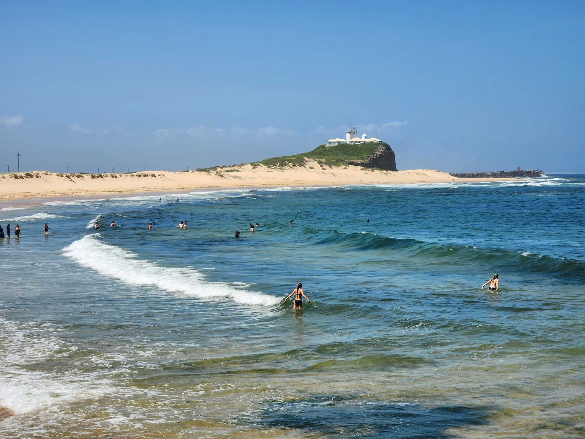 A scenic beach scene at Newcastle, New South Wales, Australia, featuring swimmers and surfers enjoying the clear blue ocean waters with moderate waves. The distinctive headland with its lighthouse and buildings is visible in the background, characteristic of Newcastle's iconic coastal landscape.