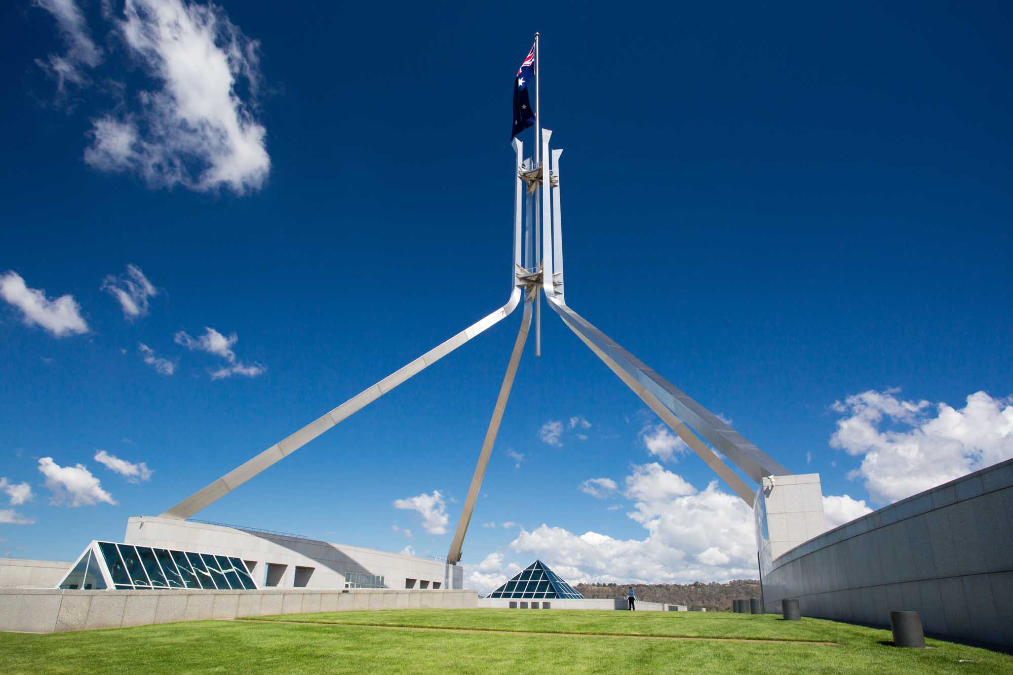 The iconic Parliament House of Australia in Canberra, Australian Capital Territory, featuring its distinctive white architecture with a tall flagpole flying the Australian flag against a brilliant blue sky. The modernist building showcases geometric design with sloping green lawns and contemporary structural elements.