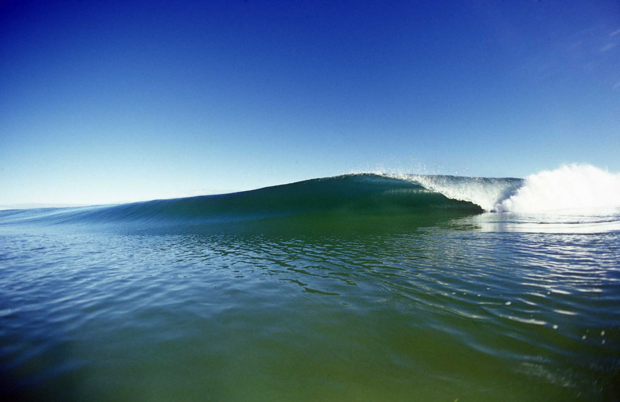 A pristine, glassy barrel wave with a smooth, cylindrical wall peeling across clear turquoise water under a bright blue sky. The wave demonstrates ideal surfing conditions with a well-defined tube and white wash breaking on the right.