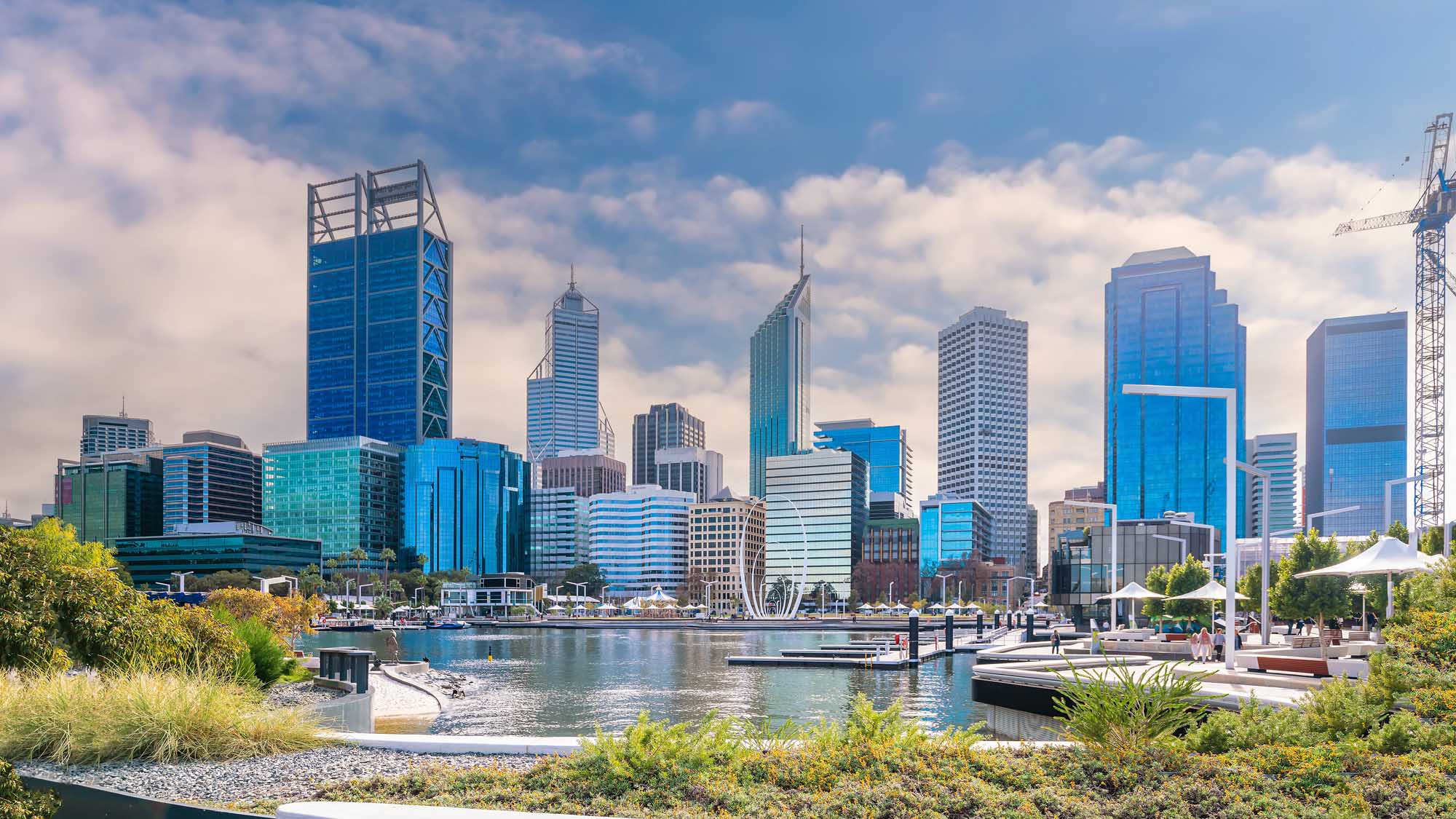 A stunning view of Perth's modern downtown skyline featuring contemporary glass and steel skyscrapers reflected in the Swan River. The image captures the city's waterfront development with landscaped riverside parks, boat docks, and a vibrant blue sky dotted with white clouds.