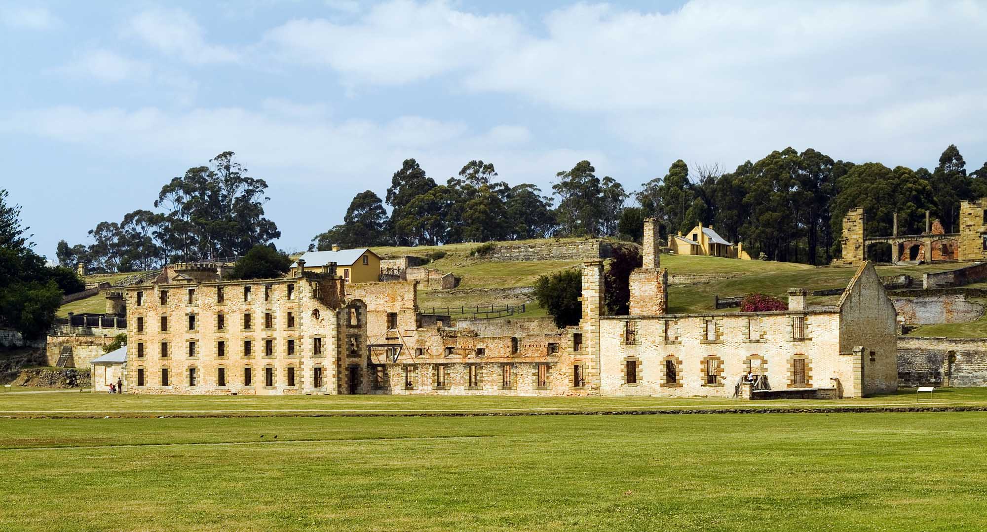 A panoramic view of Port Arthur's iconic convict-era stone buildings, featuring the large multi-story penitentiary structure and adjacent historic ruins set within a grassy field, with eucalyptus trees and rolling hills in the background under a partly cloudy sky.