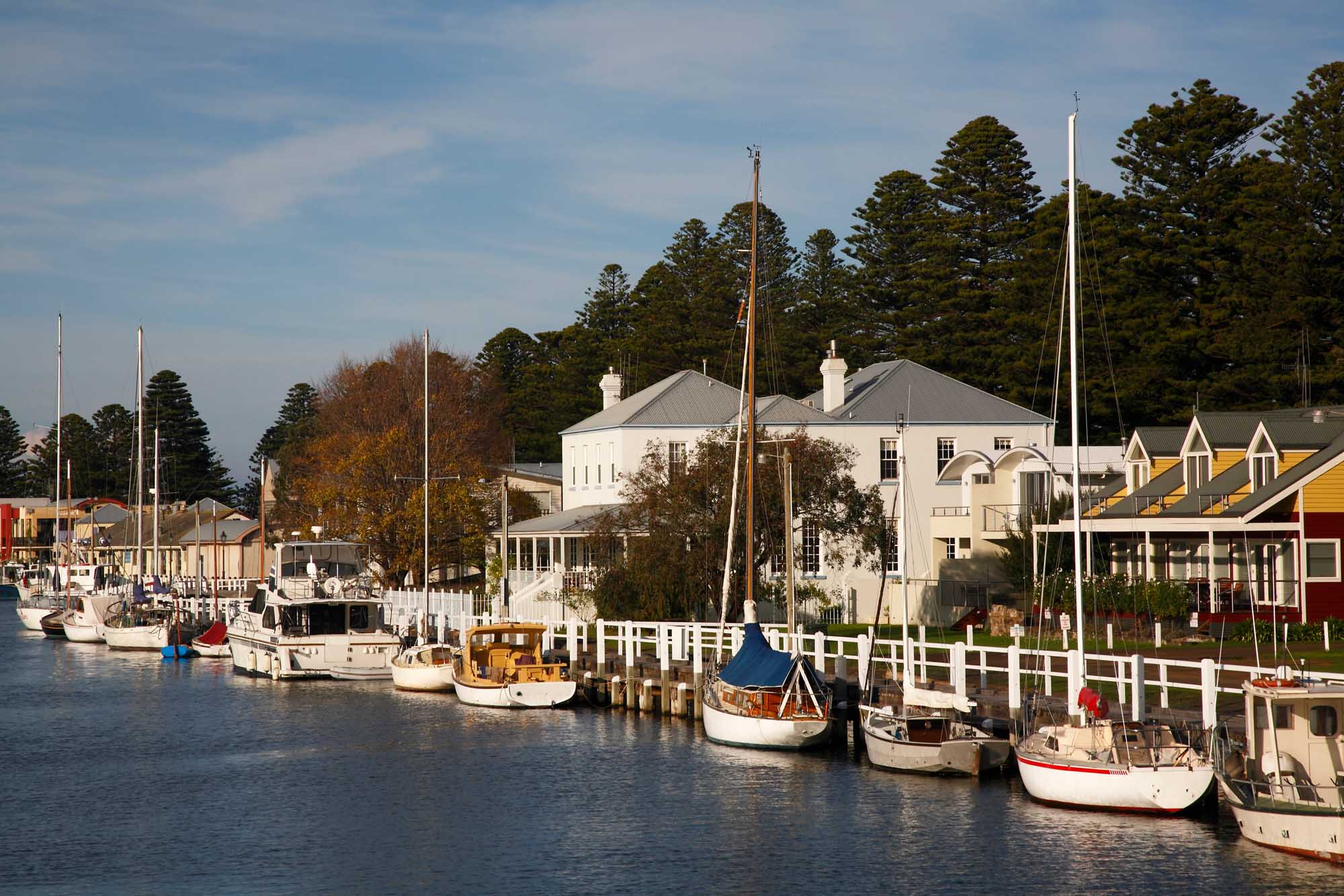 A picturesque riverside scene in Port Fairy, southwest Victoria, Australia, featuring a tranquil waterfront lined with fishing boats and pleasure craft moored at white wooden docks. Heritage and contemporary buildings, including a prominent white colonial structure, overlook the calm waters, with tall pine trees framing the scene against a clear blue sky.