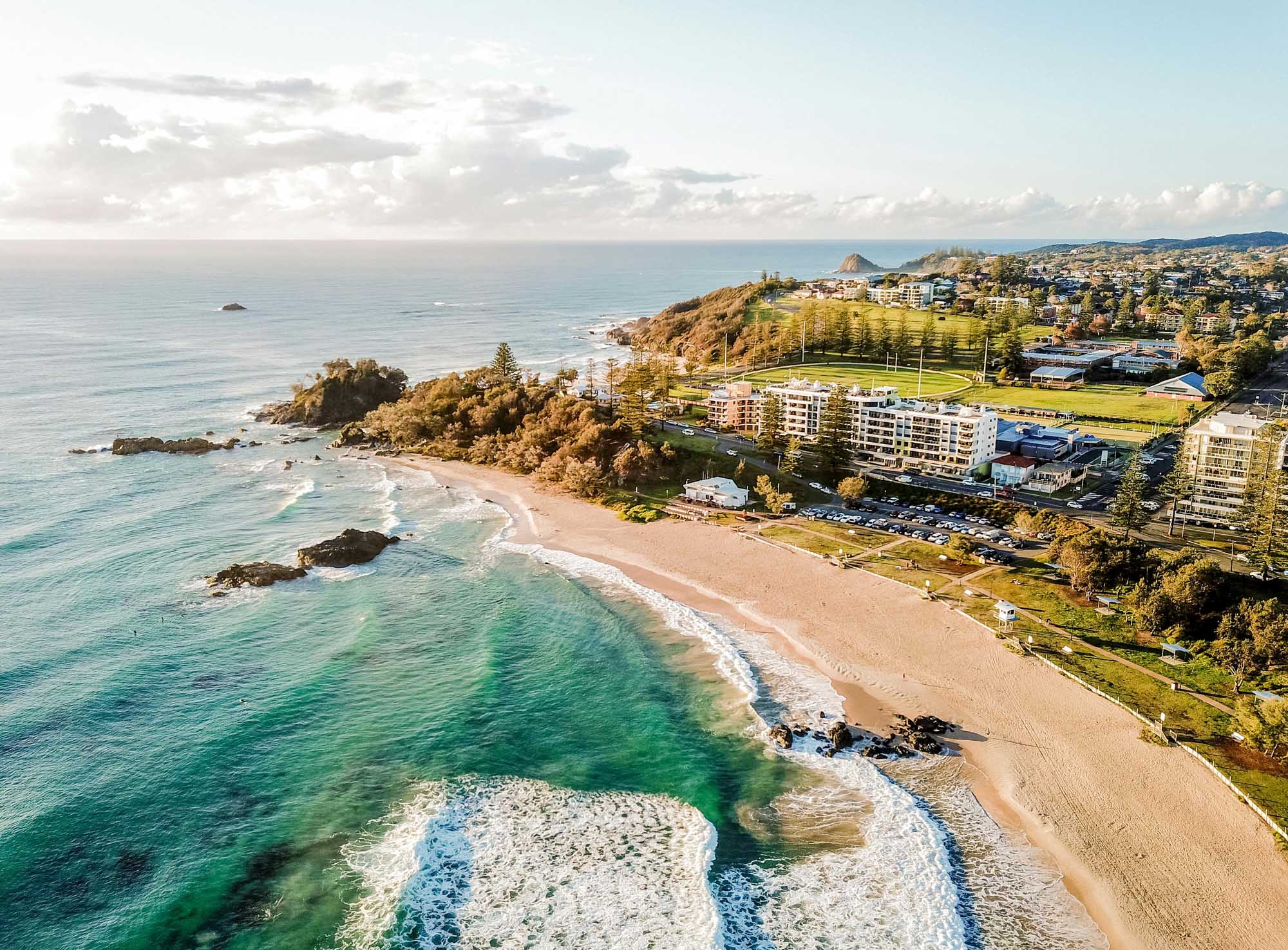 An aerial view of a scenic Australian coastal town featuring a pristine sandy beach, turquoise waters, rocky headlands, and residential buildings nestled among lush green spaces and pine trees. The image captures the natural beauty of Port Macquarie's waterfront with gentle waves rolling onto the shore and distant ocean vistas.