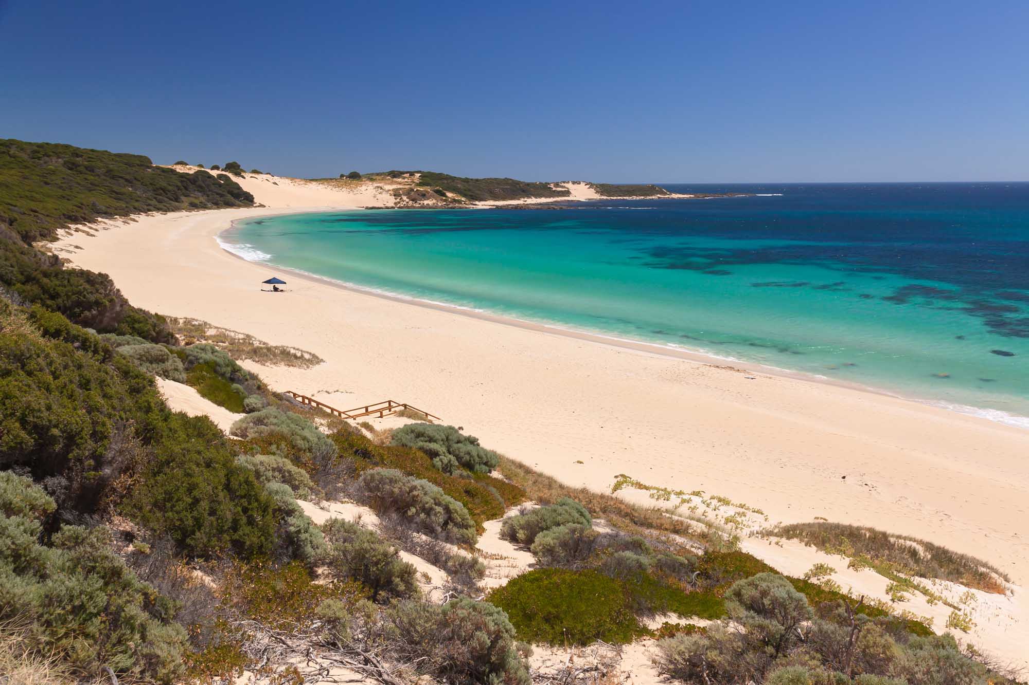 A stunning coastal landscape featuring a pristine white sand beach with crystalline turquoise waters, backed by windswept coastal dunes and native vegetation. The secluded beach is framed by dramatic coastal cliffs and offers pristine swimming conditions under clear blue skies.