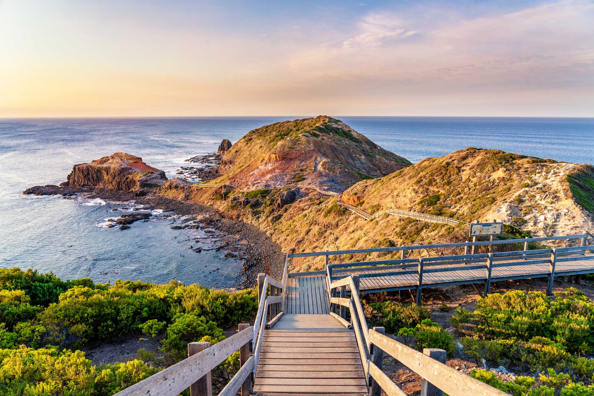 A scenic wooden boardwalk winds through coastal vegetation toward the distinctive Pulpit Rock sea stack on the Mornington Peninsula, with golden-hour sunlight illuminating the rugged volcanic formations and surrounding ocean landscape.
