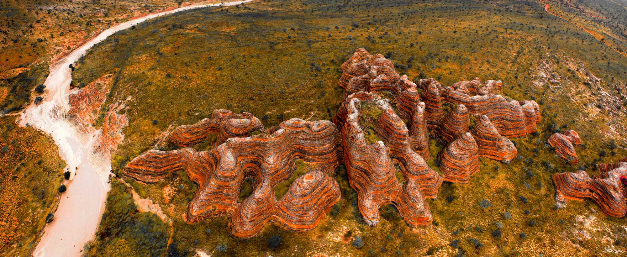 An aerial view of the distinctive beehive-shaped rock formations in Purnululu National Park (The Bungle Bungle), Western Australia, with a winding river cutting through the orange and rust-colored striped domes set against green vegetation and pale mineral deposits.