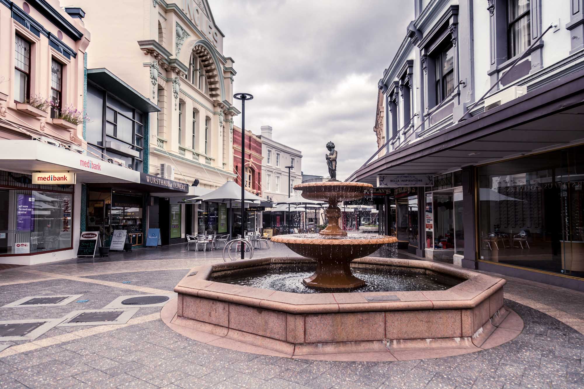 A vibrant pedestrian shopping mall in Launceston, Tasmania featuring a central tiered water fountain with bronze sculpture, surrounded by heritage-listed Victorian and Edwardian architecture. The scene captures modern retail storefronts including a Medibank branch, with cloudy skies overhead and outdoor seating areas lining the promenade.