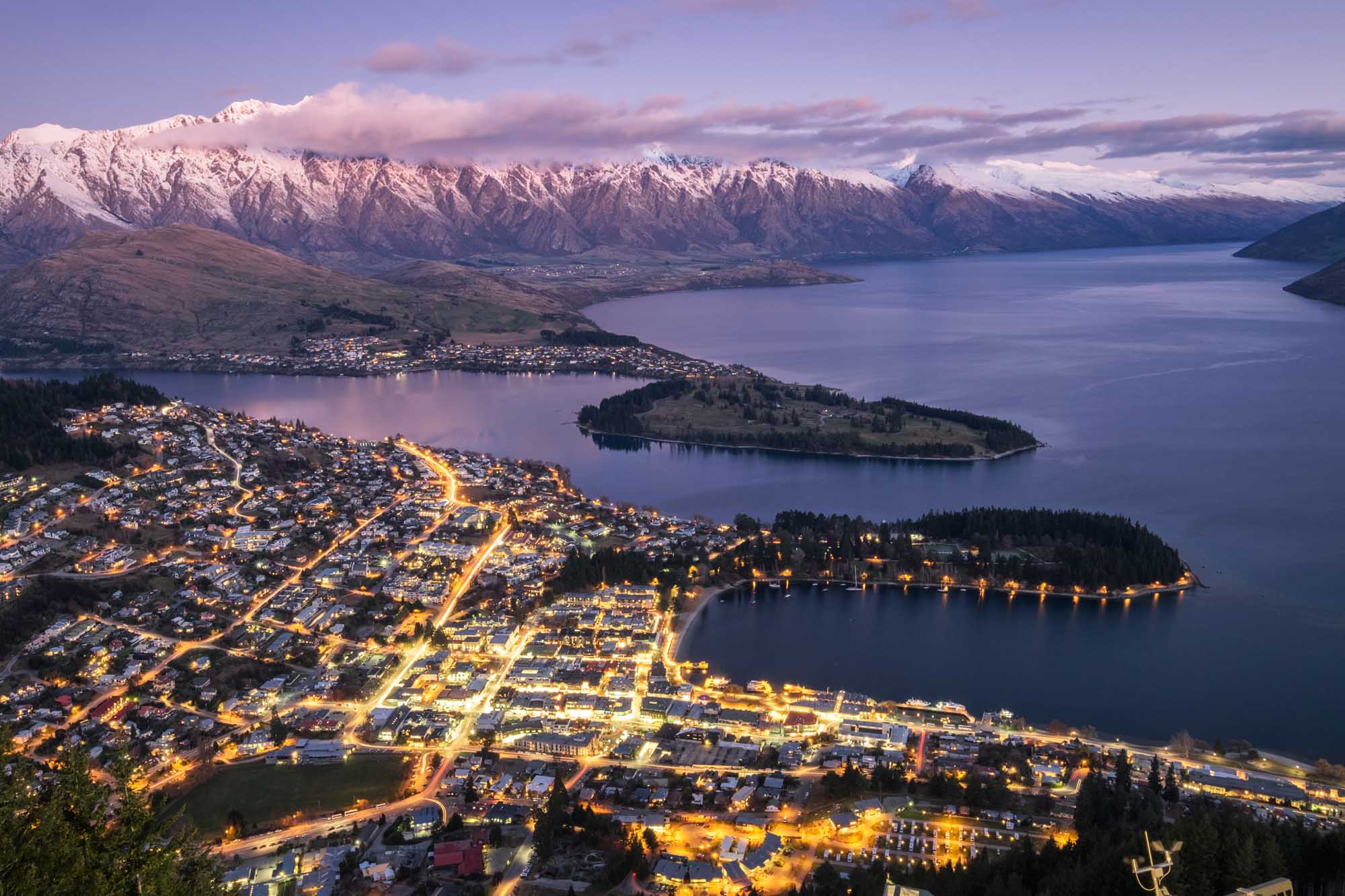 An aerial twilight photograph of Queenstown, New Zealand, showcasing the illuminated city nestled between Lake Wakatipu and surrounding mountain ranges with snow-capped peaks. The image captures the town's lights reflecting on the water, with a peninsula jutting into the lake and distant mountains visible under a purple-pink sky.