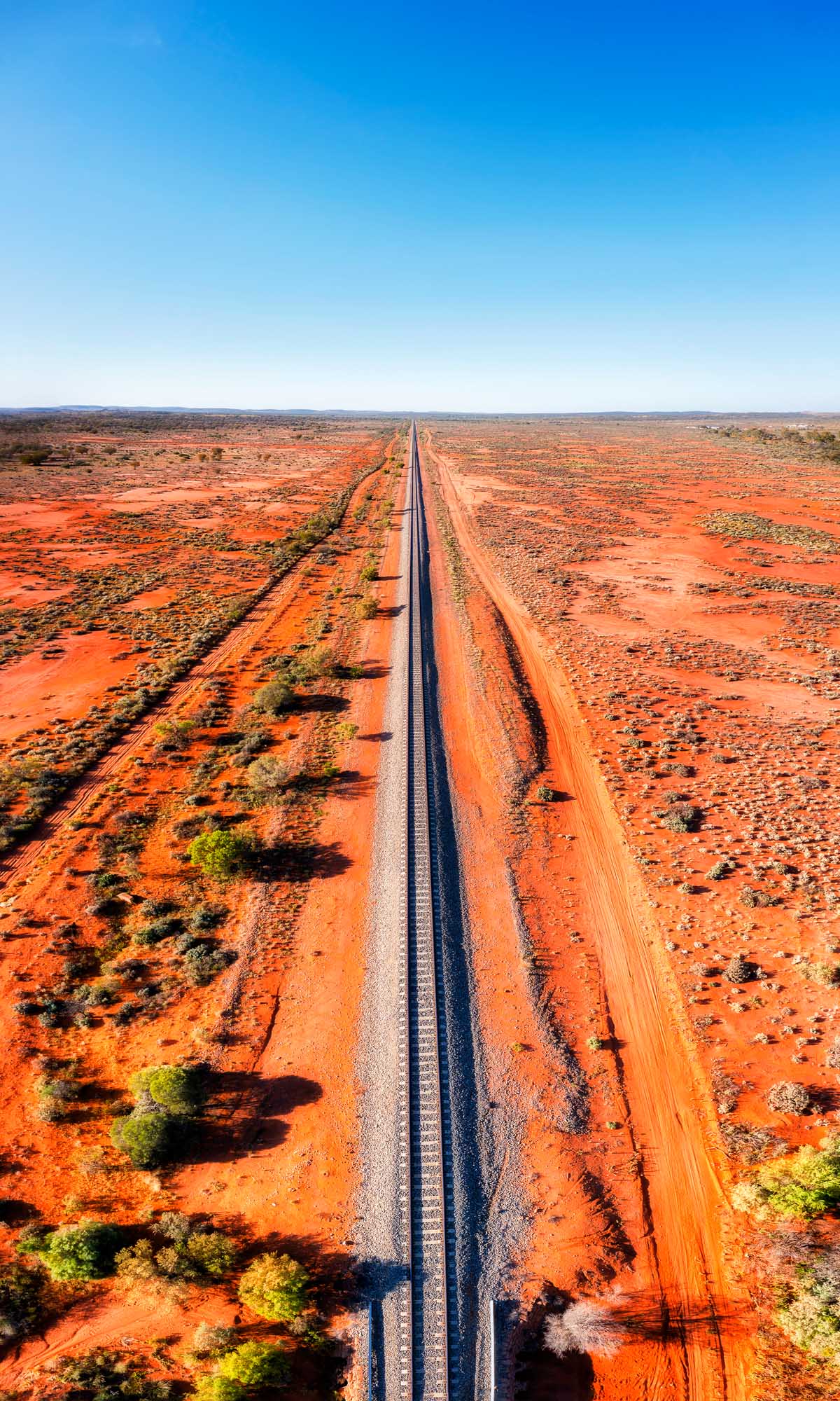 An aerial panoramic view of a railway track stretching into the vast red-soil Australian Outback, with sparse vegetation dotting the arid landscape. The image captures the iconic sight of infrastructure cutting through the remote wilderness toward Broken Hill in Far West New South Wales.