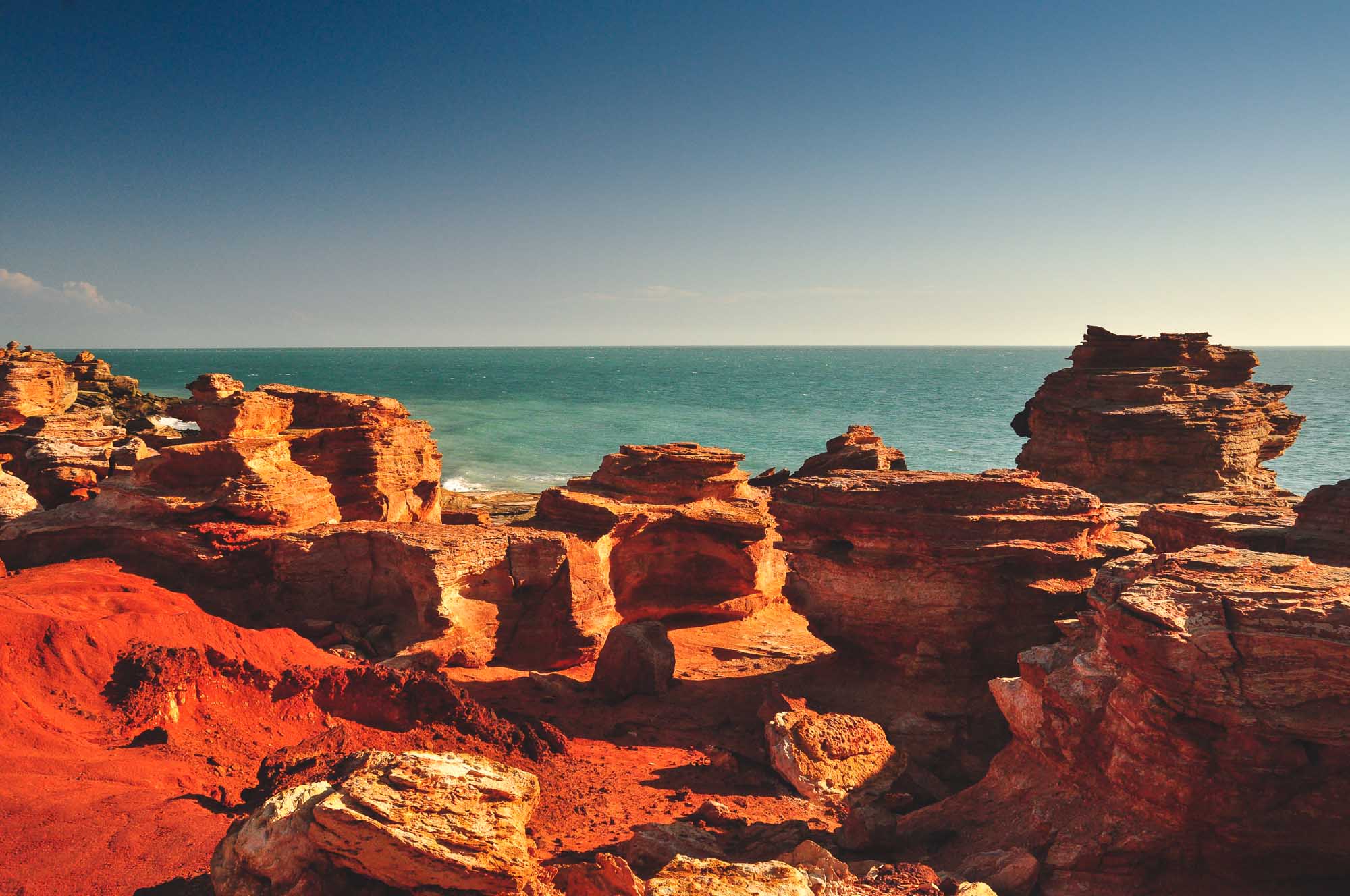 Dramatic red and orange rock formations jut from the turquoise waters at Gantheaume Point in Broome, Western Australia. The distinctive layered sandstone cliffs are illuminated by golden sunlight, creating striking contrasts against the clear blue sky and pristine ocean waters.