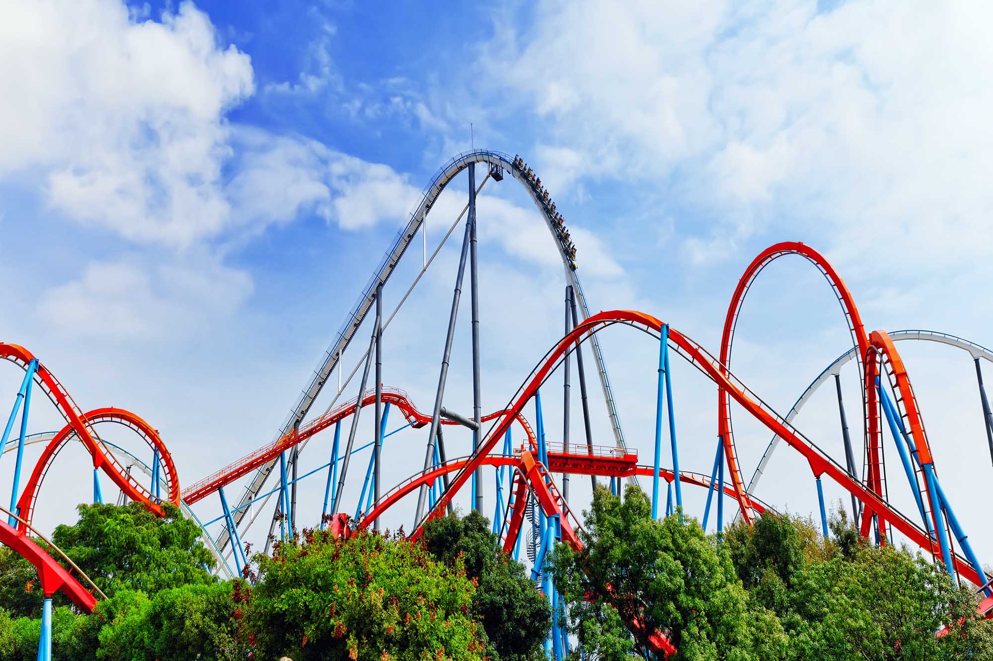 A vibrant roller coaster with striking red and blue track sections against a bright blue sky with white clouds. The ride features multiple inversions and tall support structures, with green trees visible at the base of the attraction.