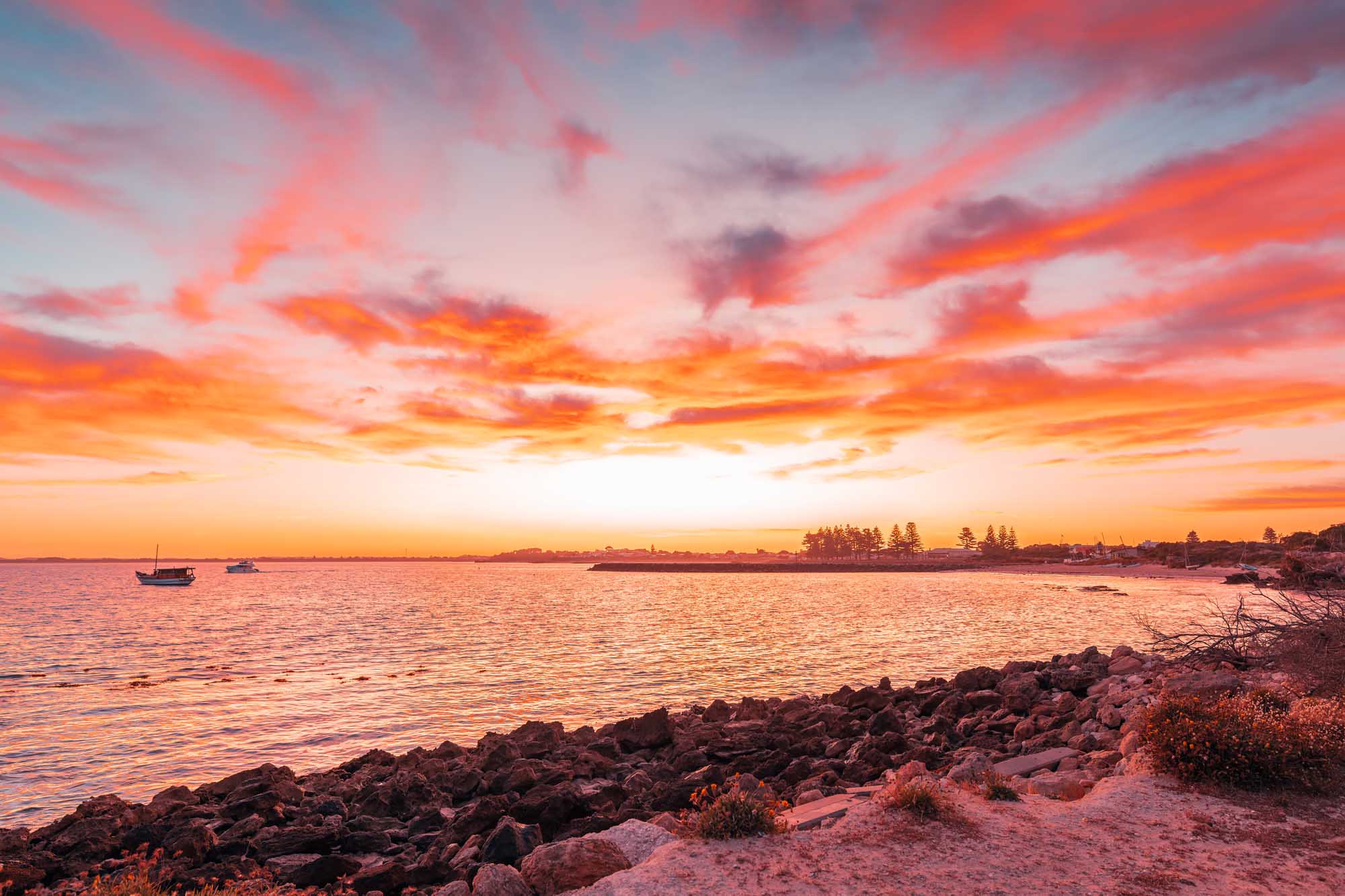 A dramatic coastal sunrise scene at Robe on South Australia's Limestone Coast, featuring moored fishing boats silhouetted against a vibrant orange and pink sky, with a rocky shoreline and sandy beach in the foreground and a tree-lined peninsula visible across the calm water.