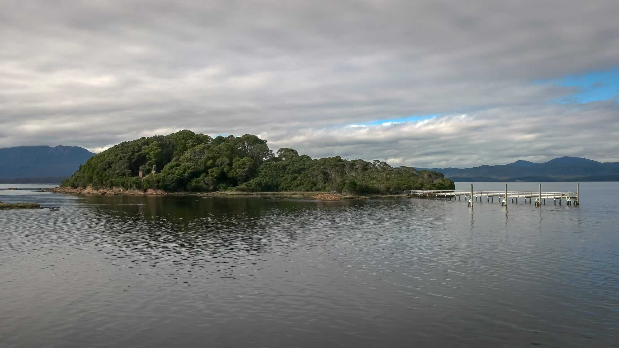 A scenic wide view of Sarah Island in Macquarie Harbour, Tasmania, featuring the historic penal settlement ruins surrounded by dense native forest and calm waters, with mountains visible in the distant background under an overcast sky.