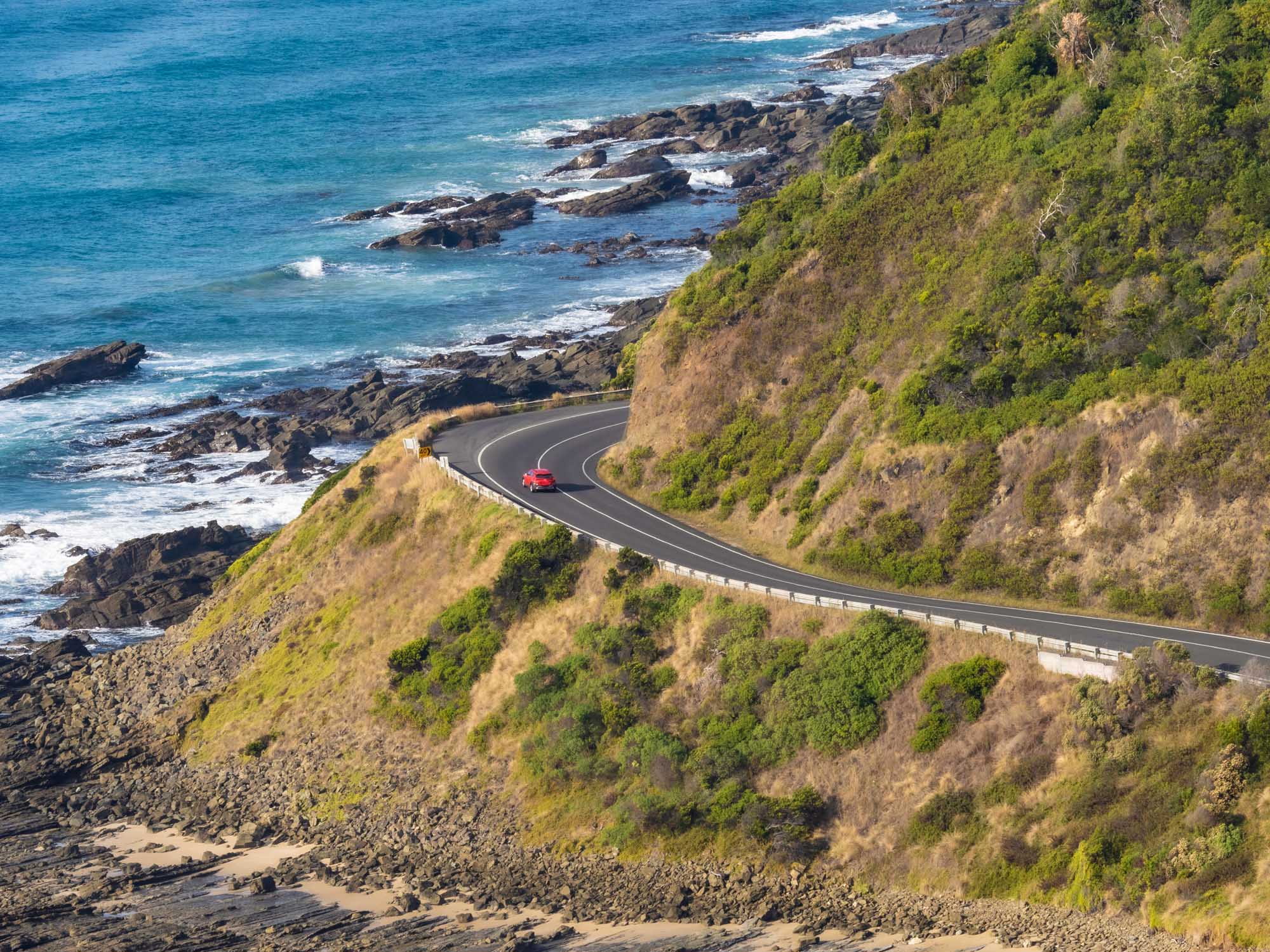 A red car navigates a winding coastal highway carved into a lush, vegetation-covered cliff overlooking a dramatic rocky shoreline with turquoise waters and white-capped waves. The road features smooth asphalt with clear lane markings and a safety barrier, set against a pristine natural seascape.
